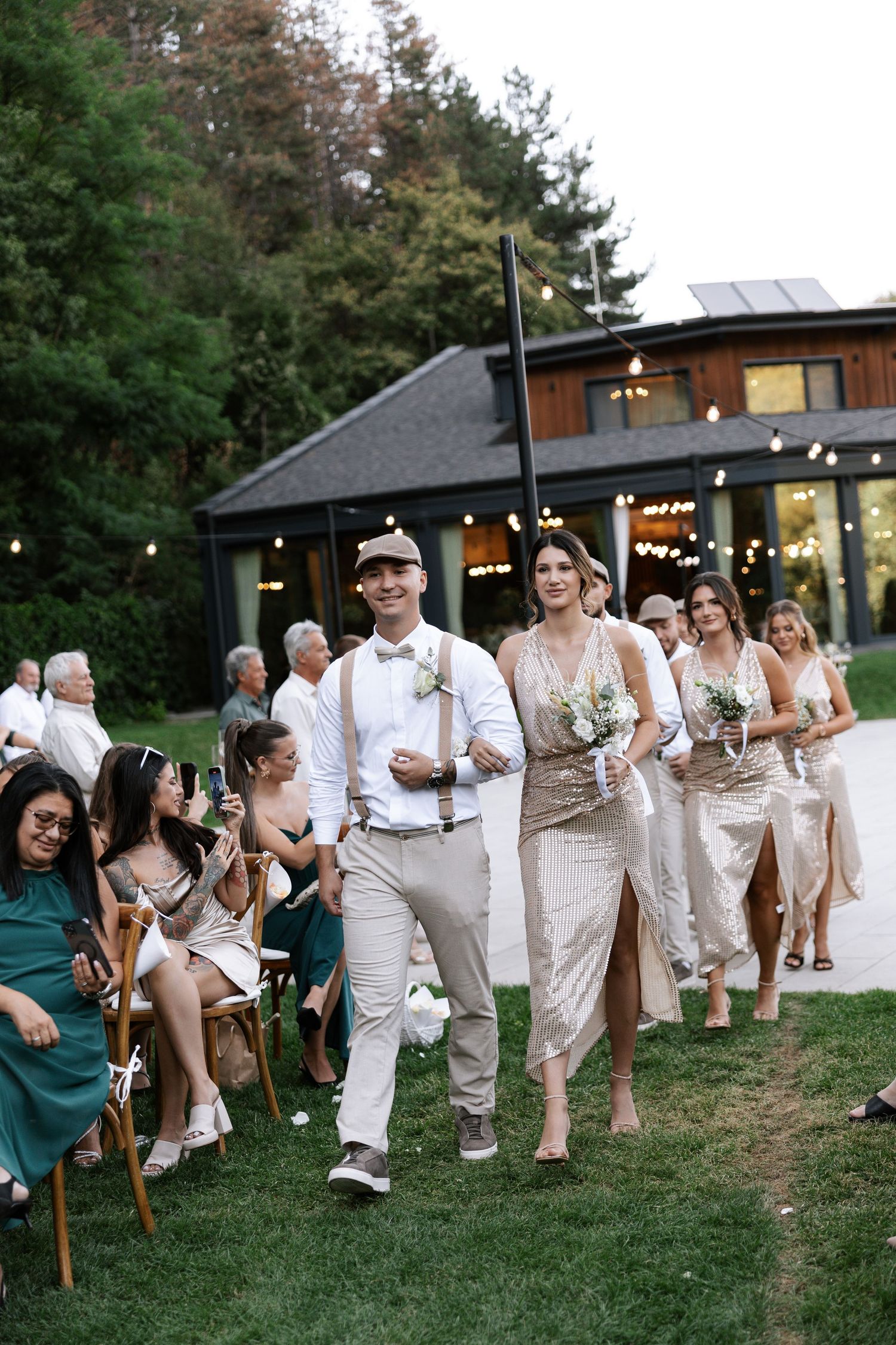 Wedding party members in coordinated white and beige outfits dance and celebrate on a grassy lawn at an outdoor venue.