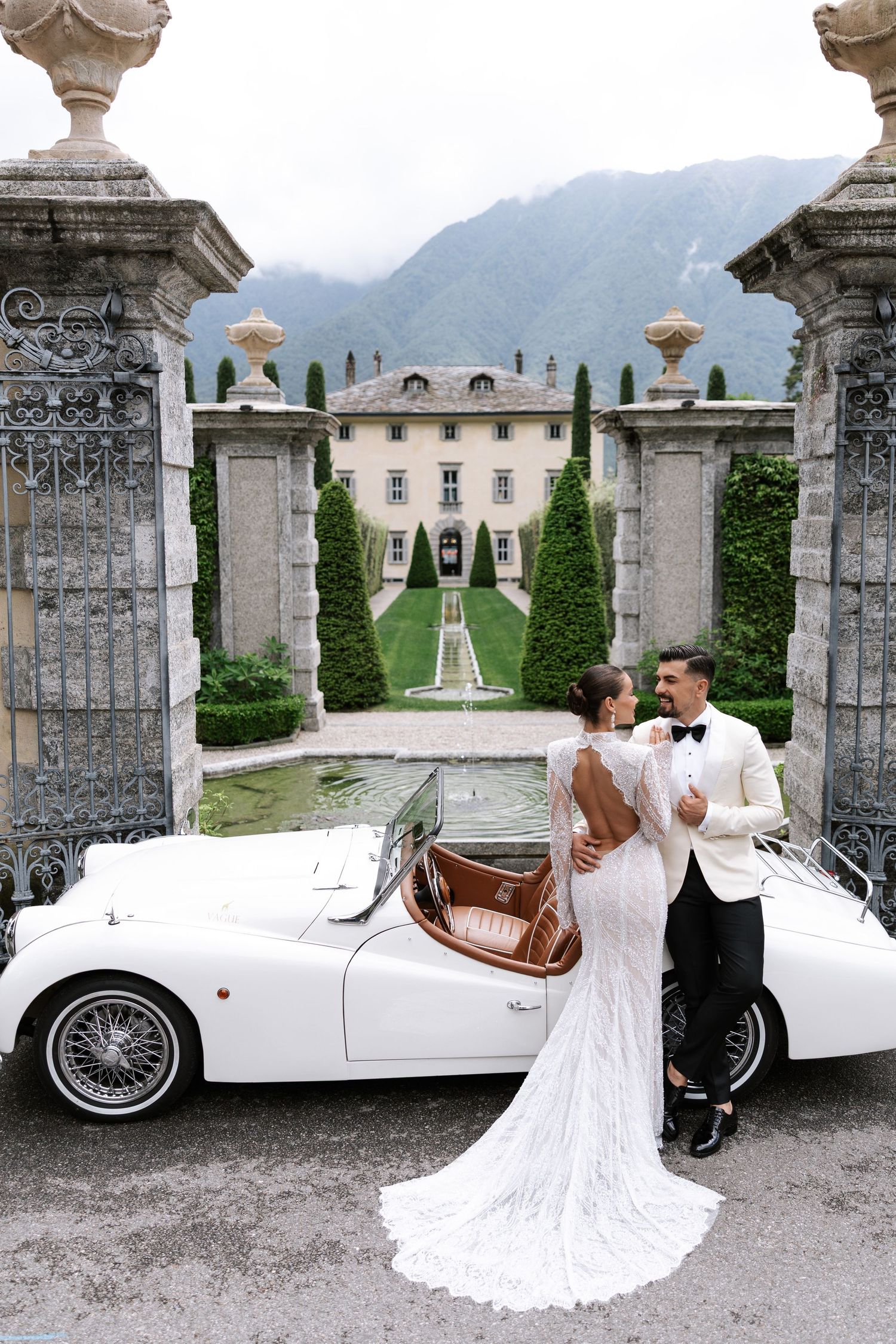 Elegant wedding couple poses with vintage white convertible car at grand Italian villa entrance with mountain backdrop.