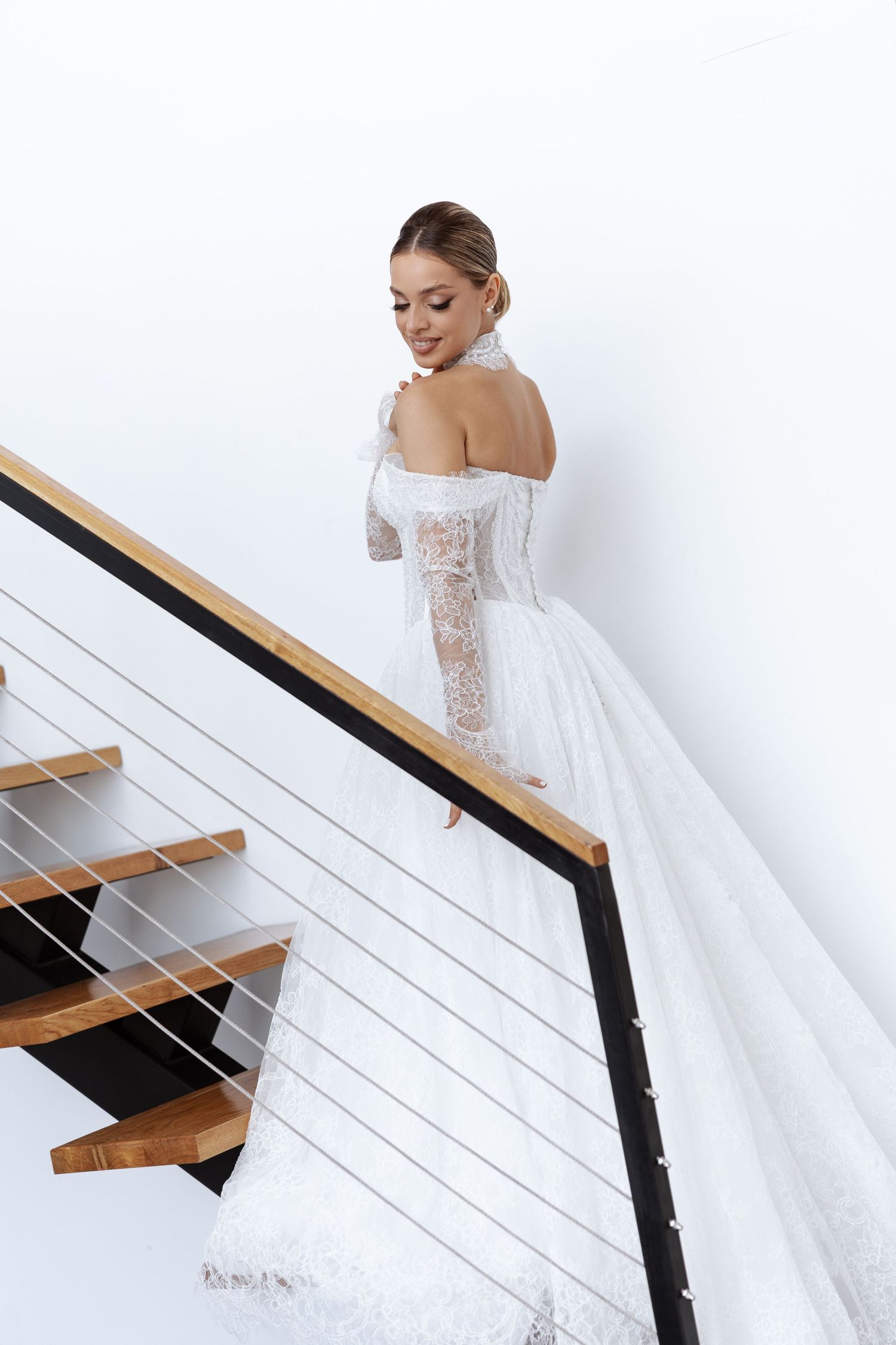 Bride in elegant lace wedding gown poses on modern staircase with wooden steps.