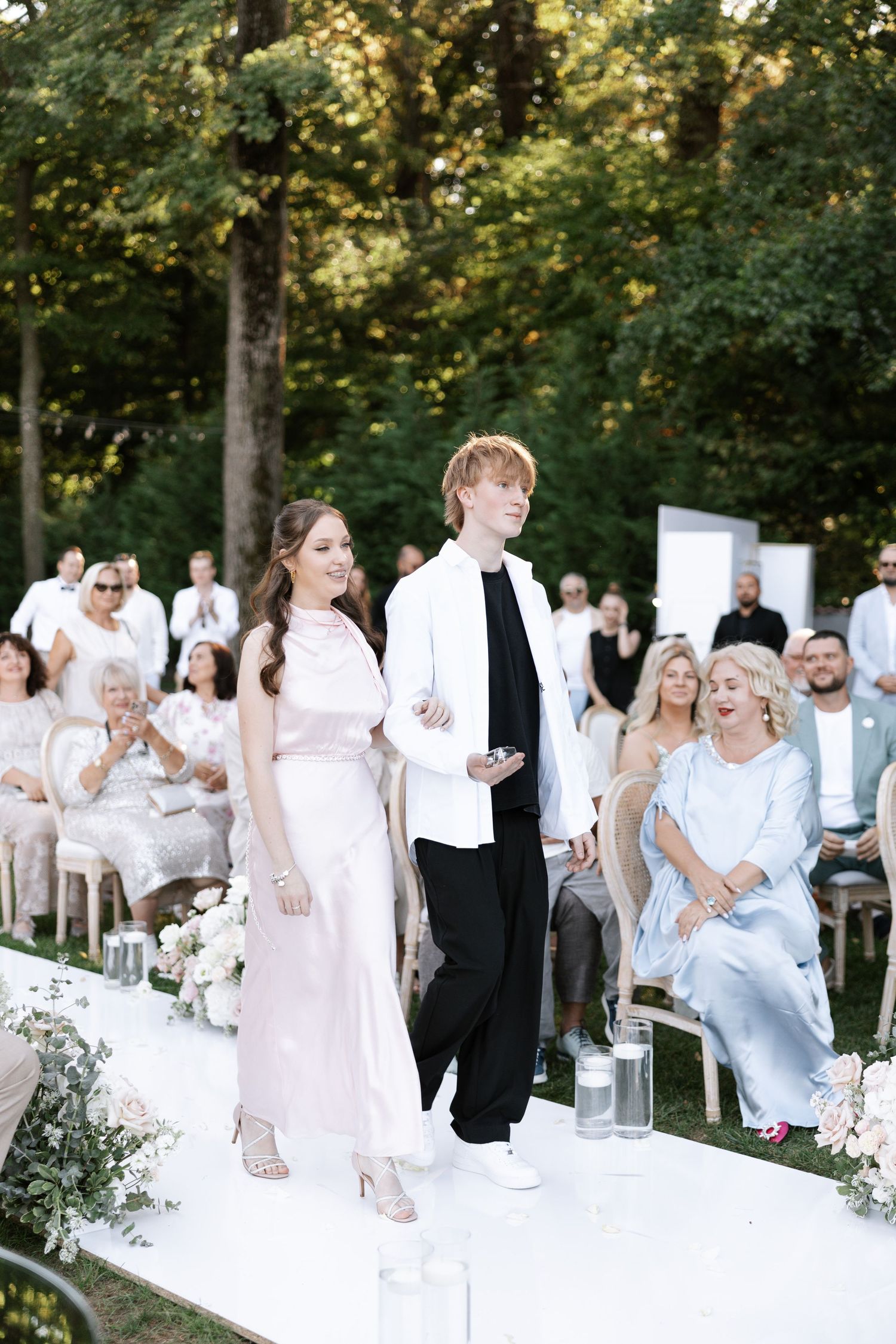 Wedding party members walking down white aisle runner during outdoor ceremony.