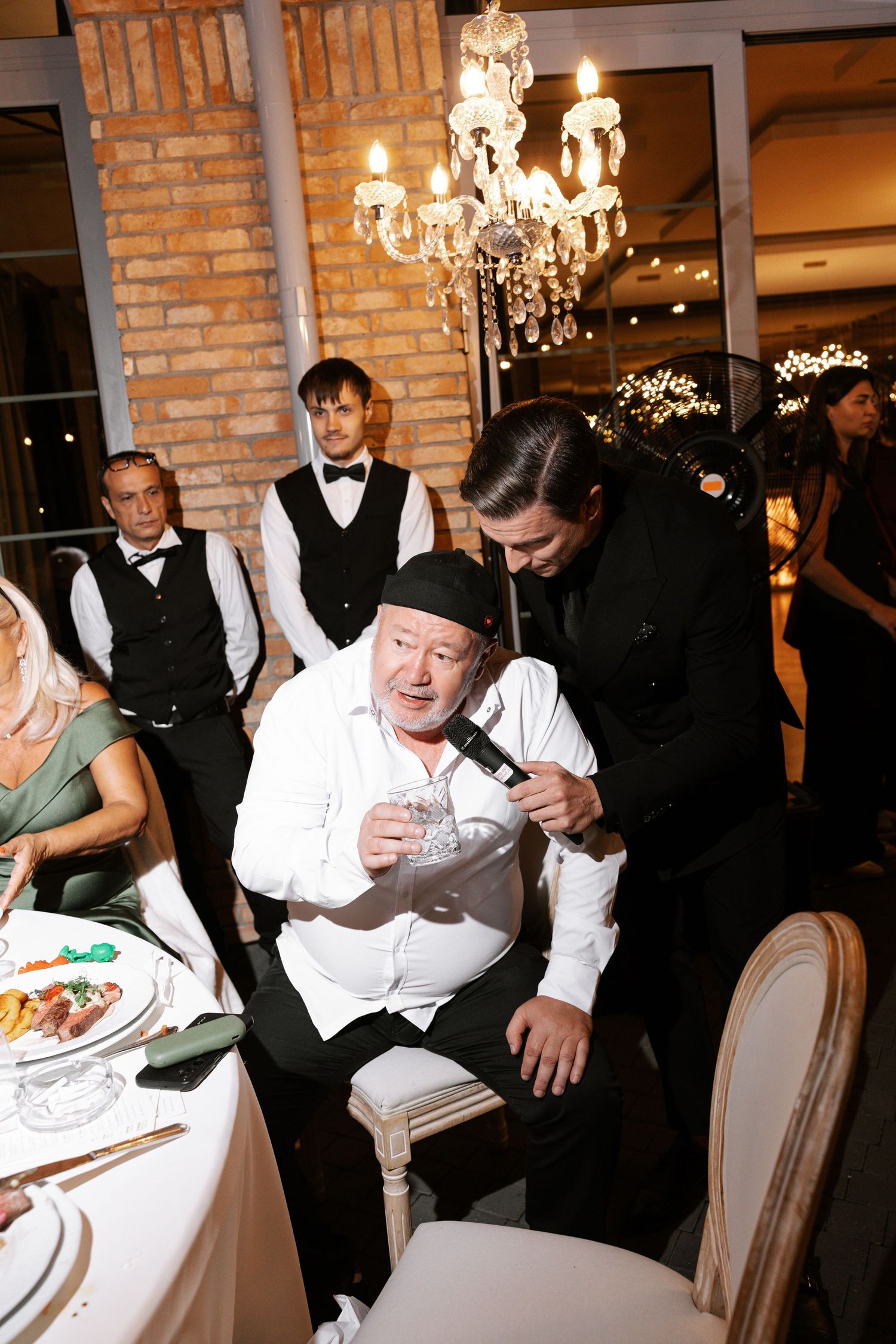 Wedding guests in formal attire seated at tables with elegant chandeliers and exposed brick walls.