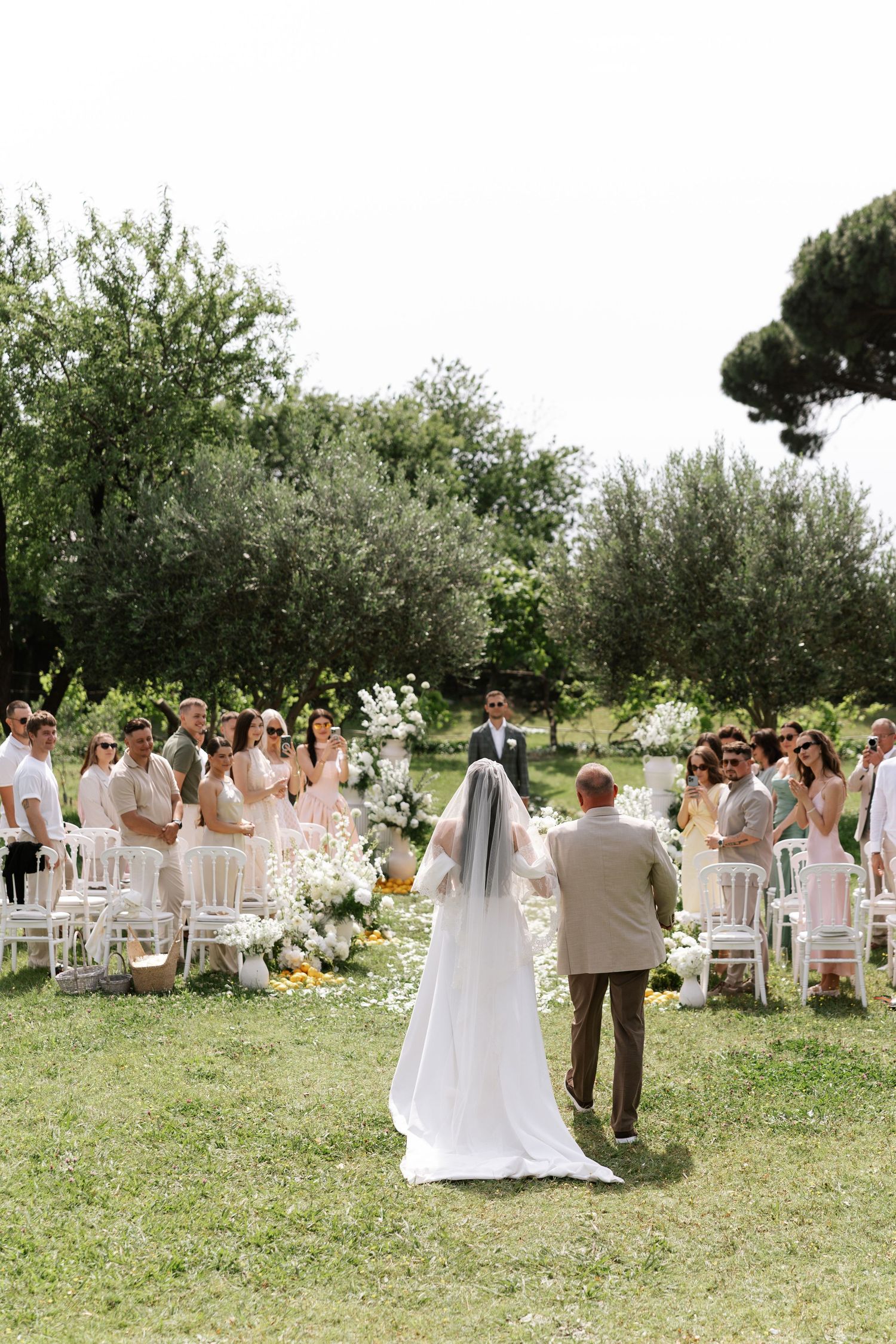 Outdoor garden wedding ceremony with white chairs arranged on grass surrounded by olive trees and natural scenery.