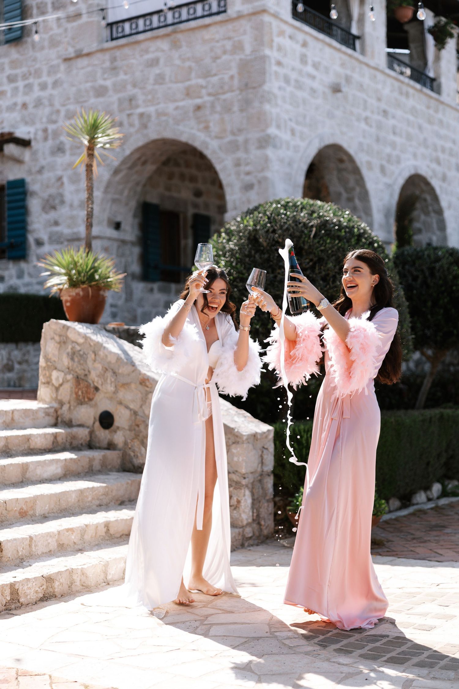 Two women in flowing pink dresses celebrating with champagne on stone steps of a Mediterranean villa.