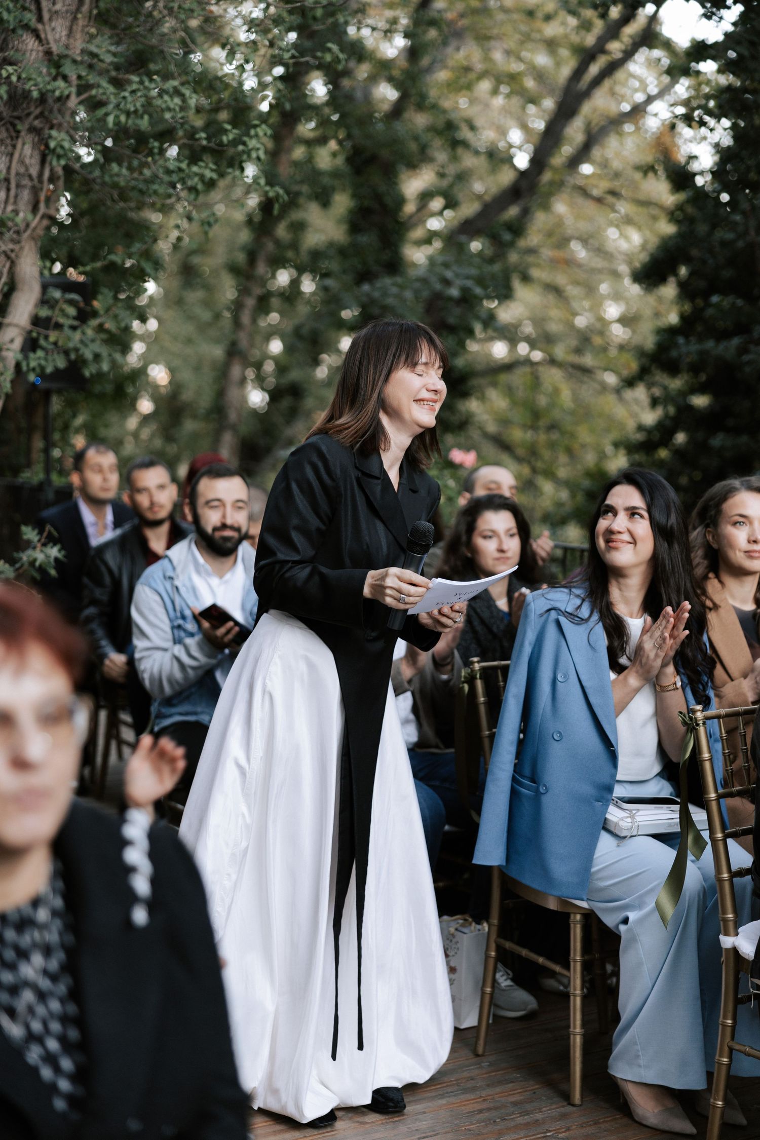 A wedding ceremony speaker addresses guests seated on wooden chairs outdoors.