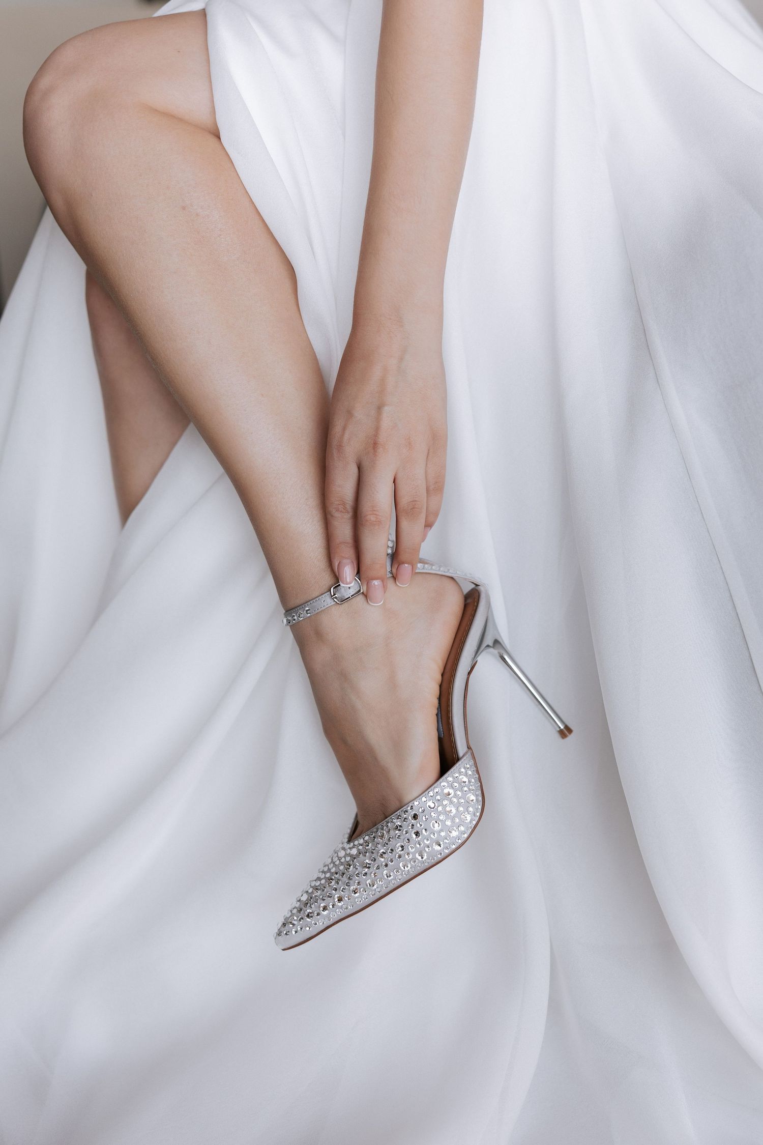 Elegant sparkly wedding heel and ankle bracelet on bride's foot against white dress fabric.