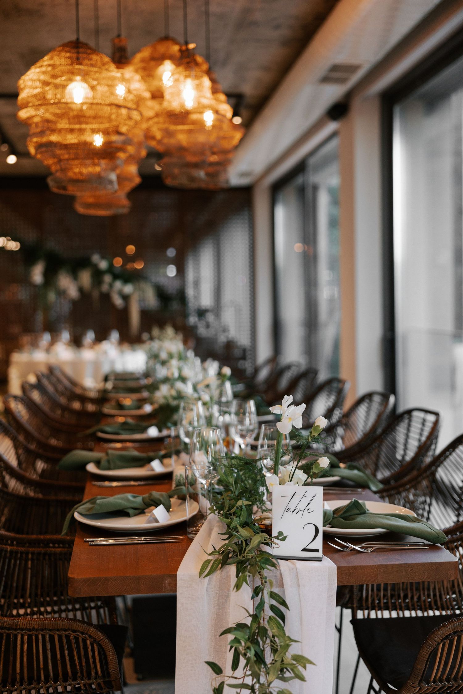 Long dining table set with place settings, green foliage runners and woven amber pendant lights above.