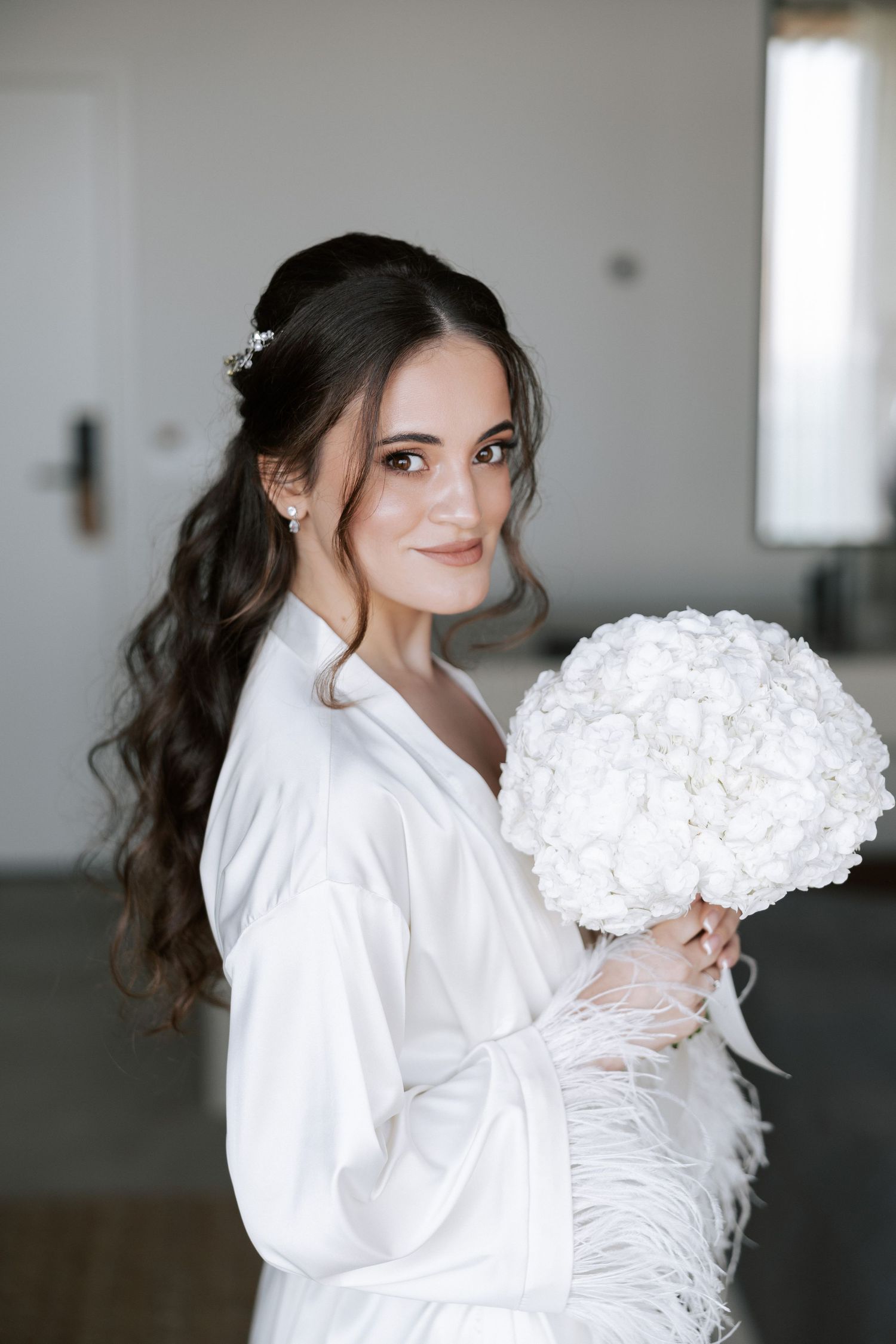 A bride in white robe holds a white hydrangea bouquet during wedding preparations.