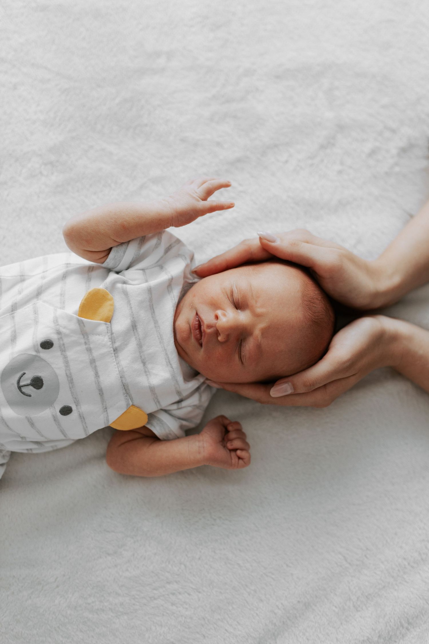 A peaceful sleeping newborn baby wearing a gray onesie with bear design lying on white bedding.