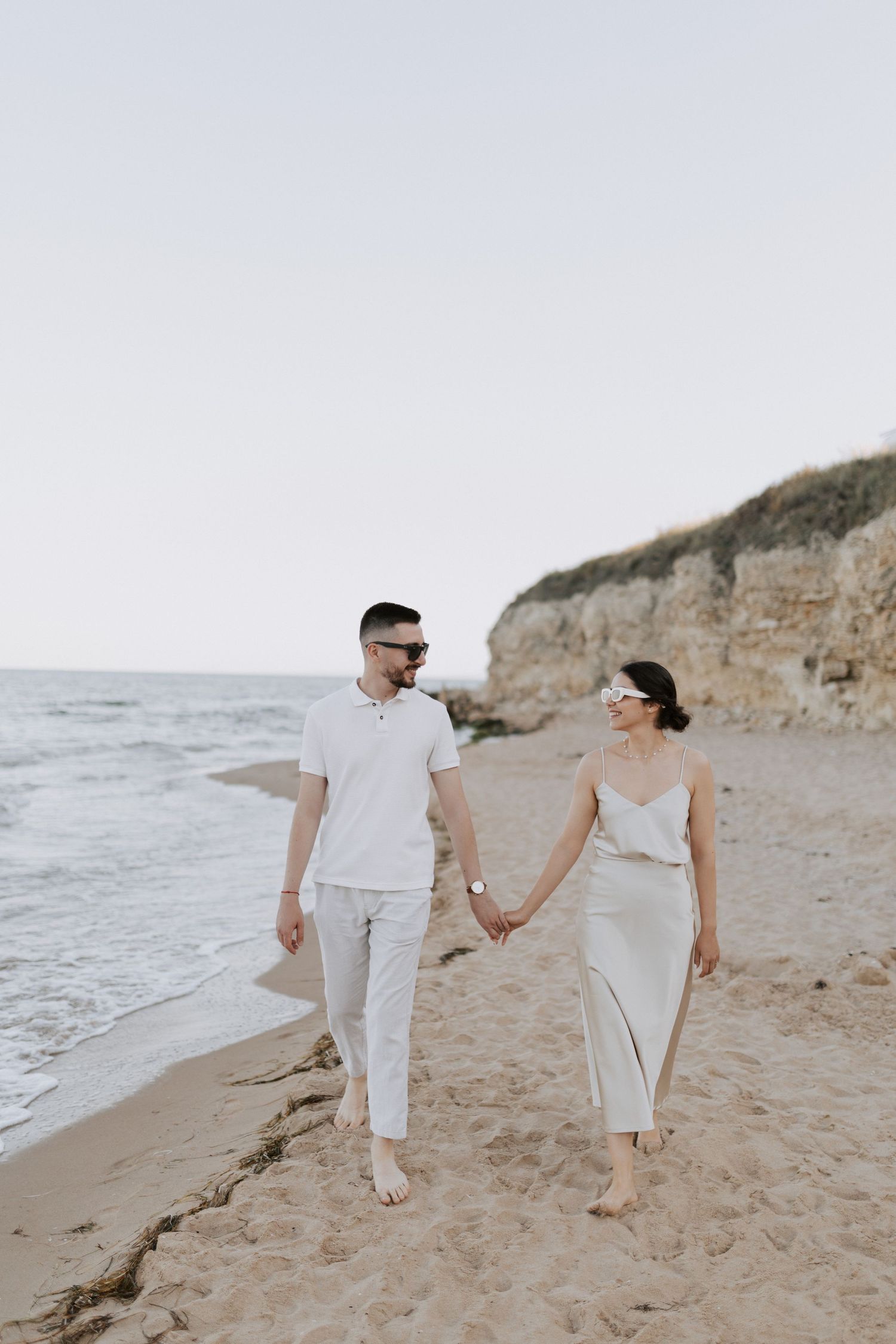 A couple holds hands while walking barefoot along a scenic beach with limestone cliffs.