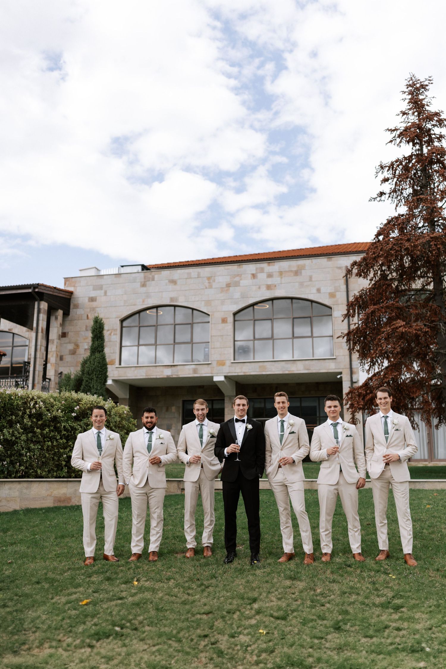 Wedding party in cream suits poses with groom in black tuxedo outside stone building.