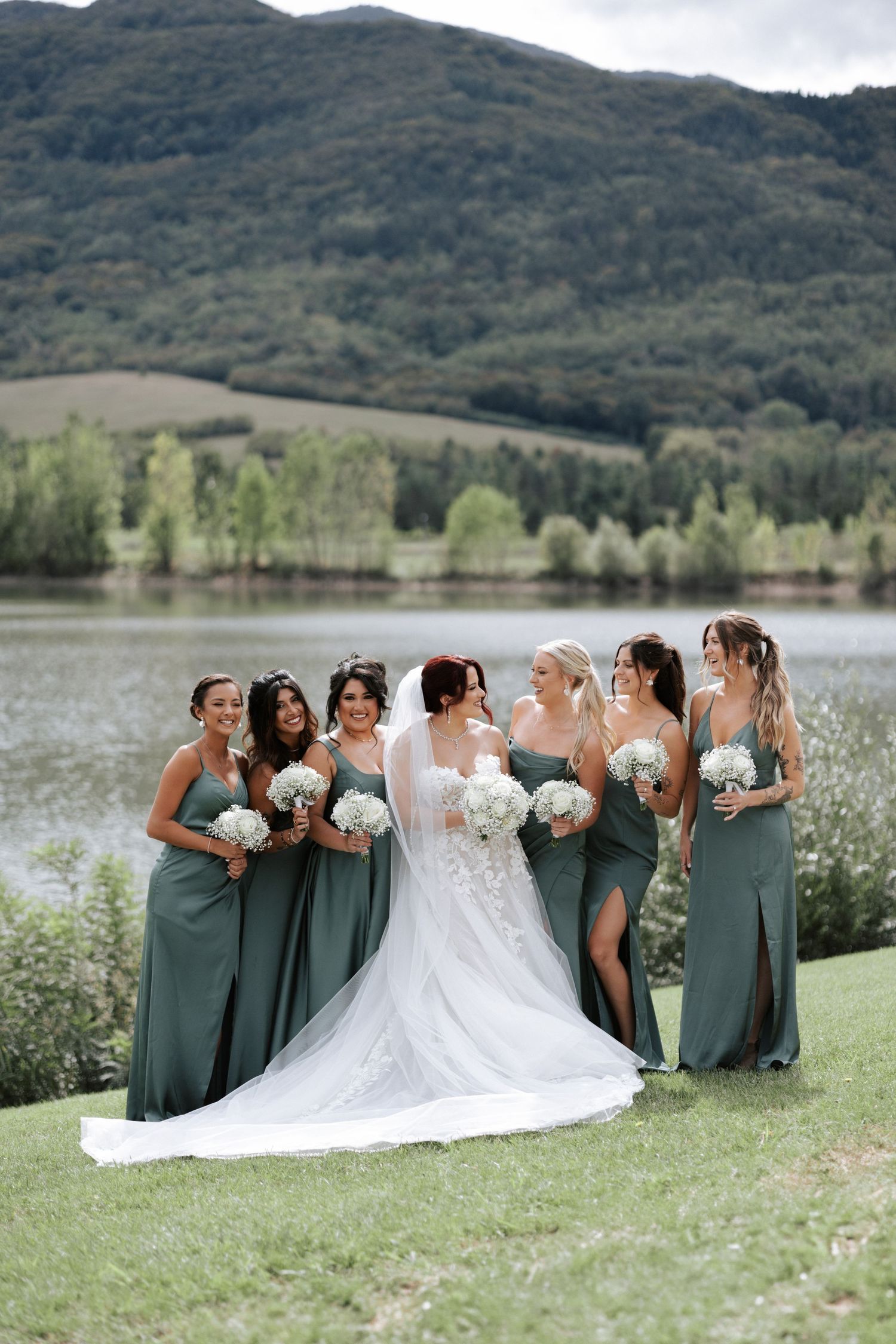 Candid moment of bridal party sharing laughter on grassy hillside by mountain lake.