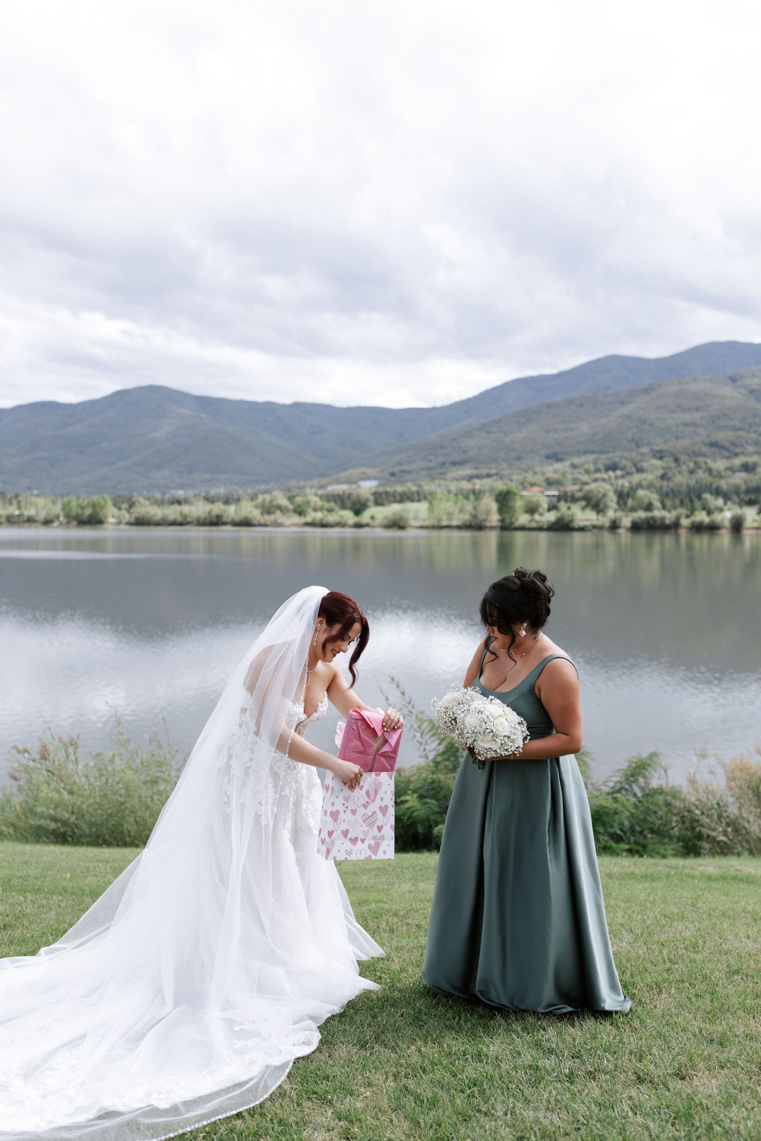 Bride in white gown and bridesmaid in sage dress look at gifts by a mountain lake.