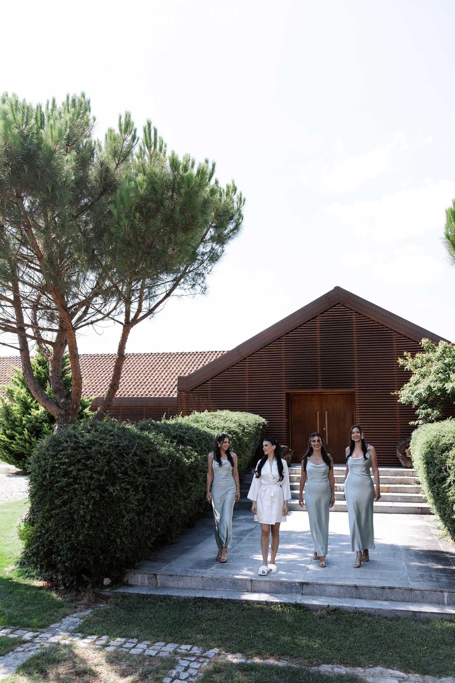 A group in white dresses walks along a path next to rustic wooden cabins and pine trees on a sunny day.