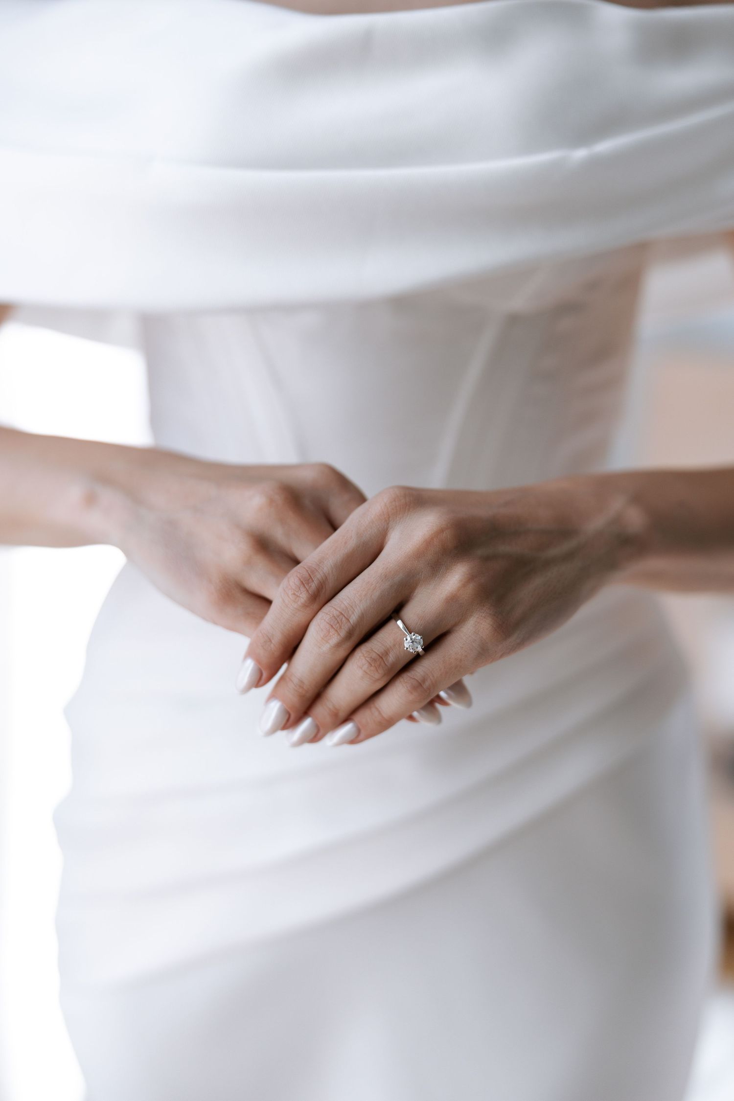 Close-up view of hands wearing an engagement ring against flowing white fabric creating an elegant bridal detail.