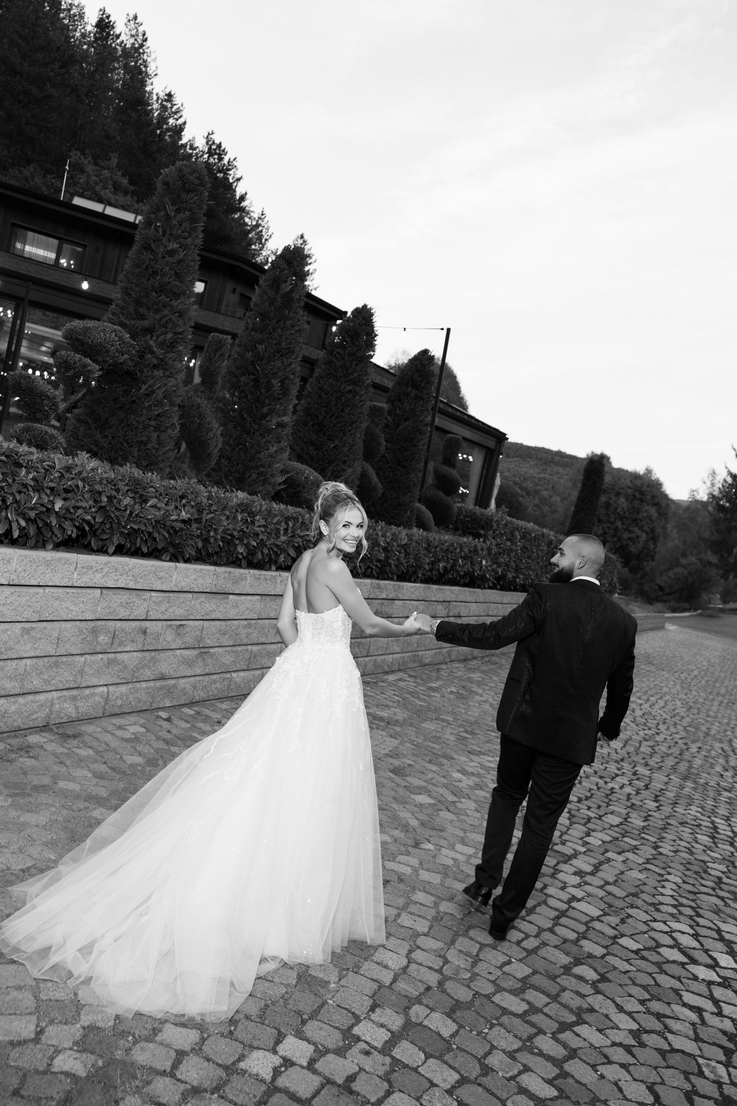 A bride in a flowing white gown and groom in black suit walk away on a cobblestone path in black and white.