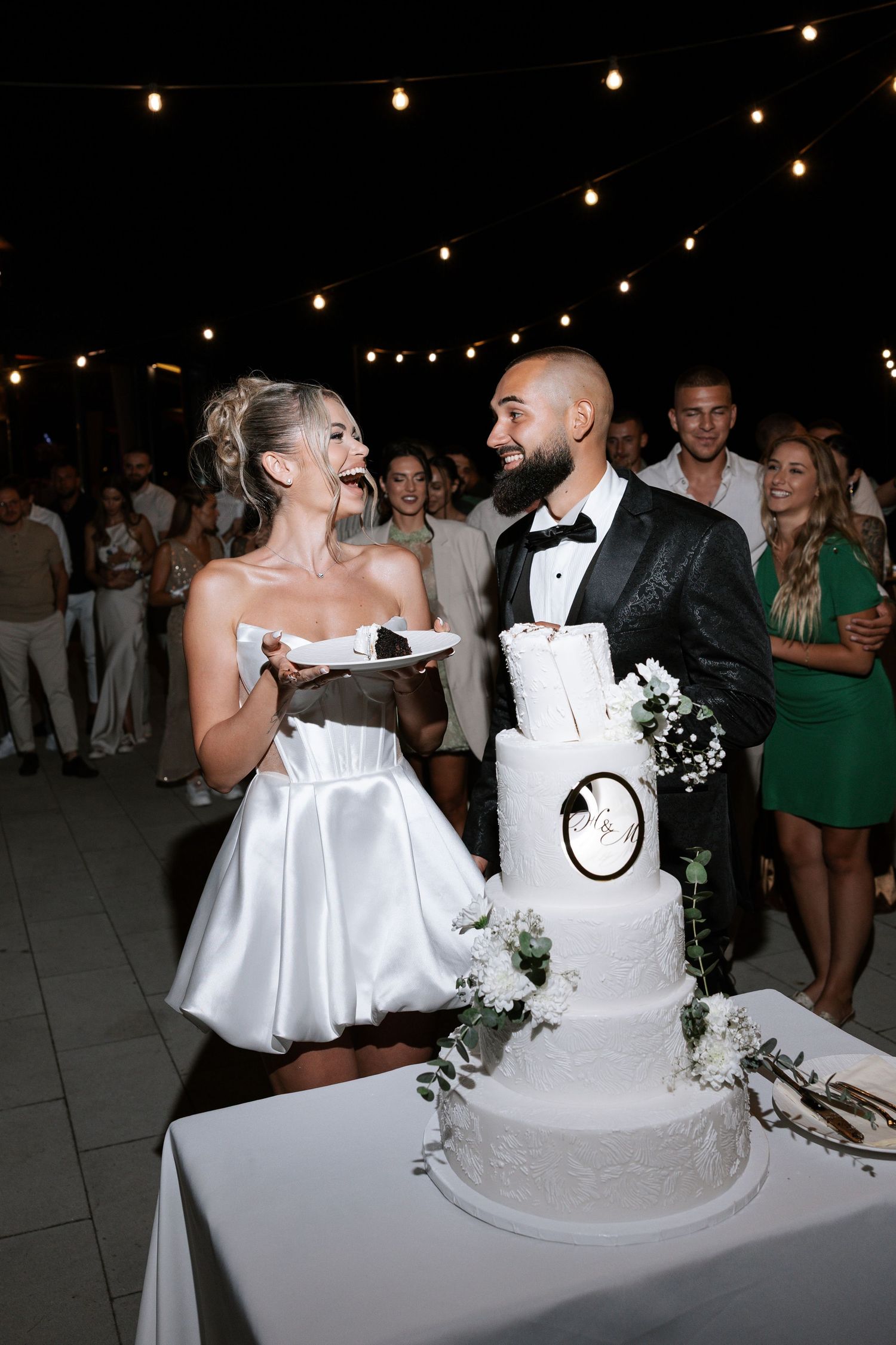 A wedding couple cuts into their elegant white tiered cake at an evening reception with string lights overhead.