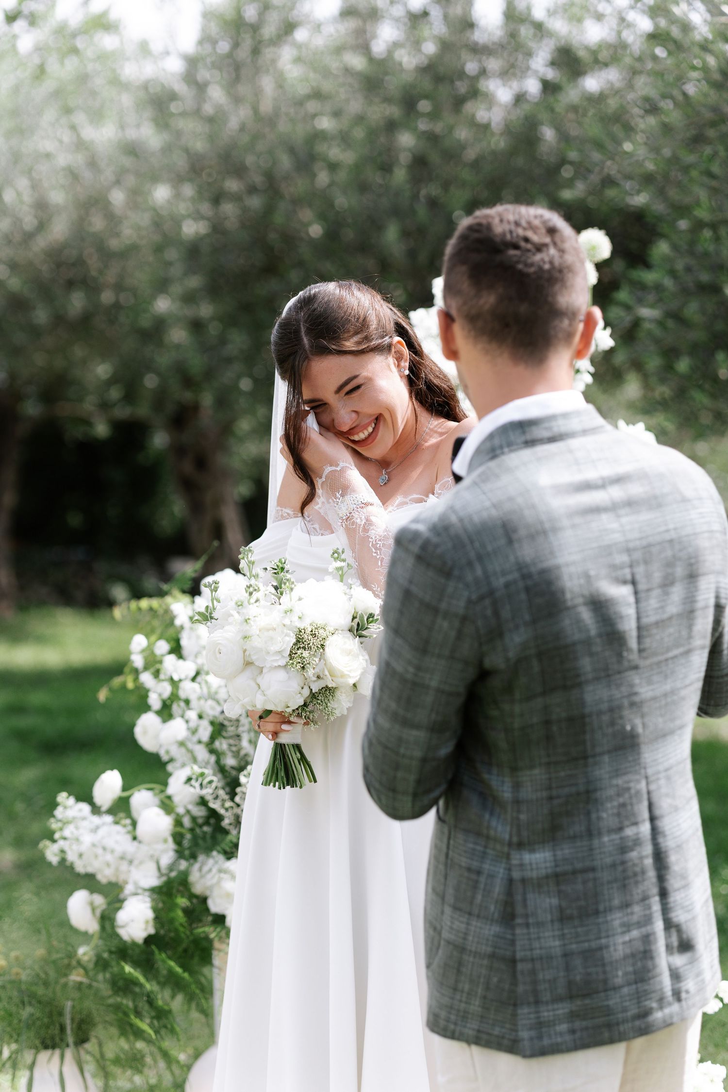 A romantic wedding couple shares joyful laughter together outdoors surrounded by greenery and white floral bouquets.