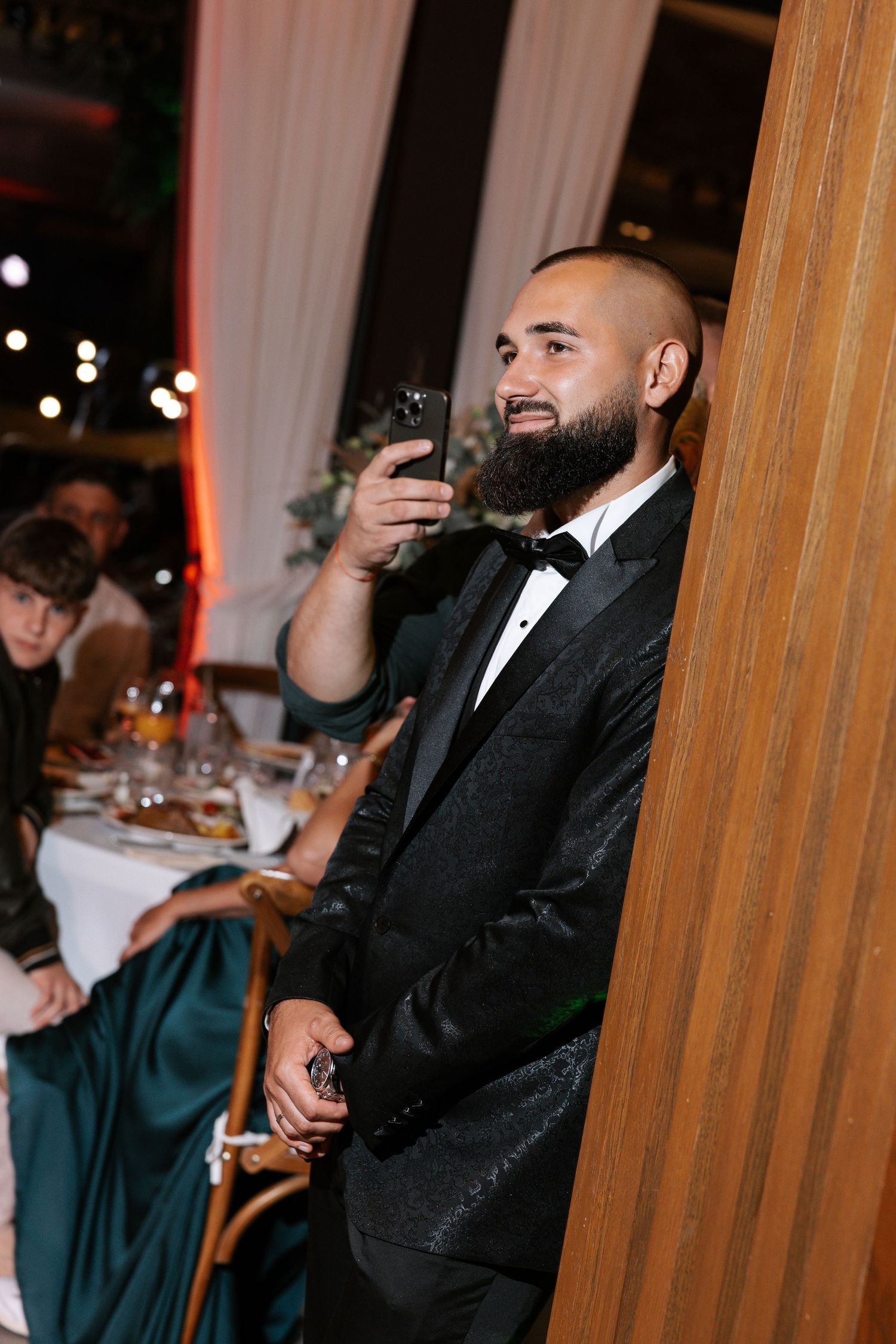 A wedding reception scene showing guests in formal attire at an elegant venue with wooden pillars and warm lighting.