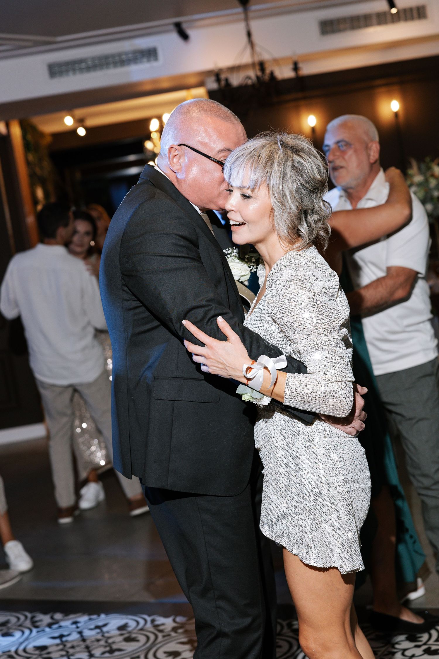 An elderly couple dances together at an event, with the woman wearing a sparkly silver top and the man in a black suit.