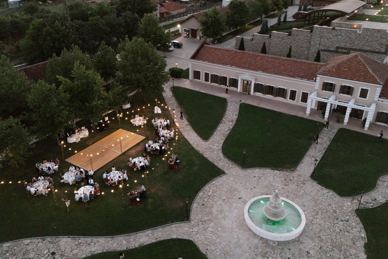 Aerial view of an outdoor wedding reception with tables set up on a lawn near a fountain and white building.