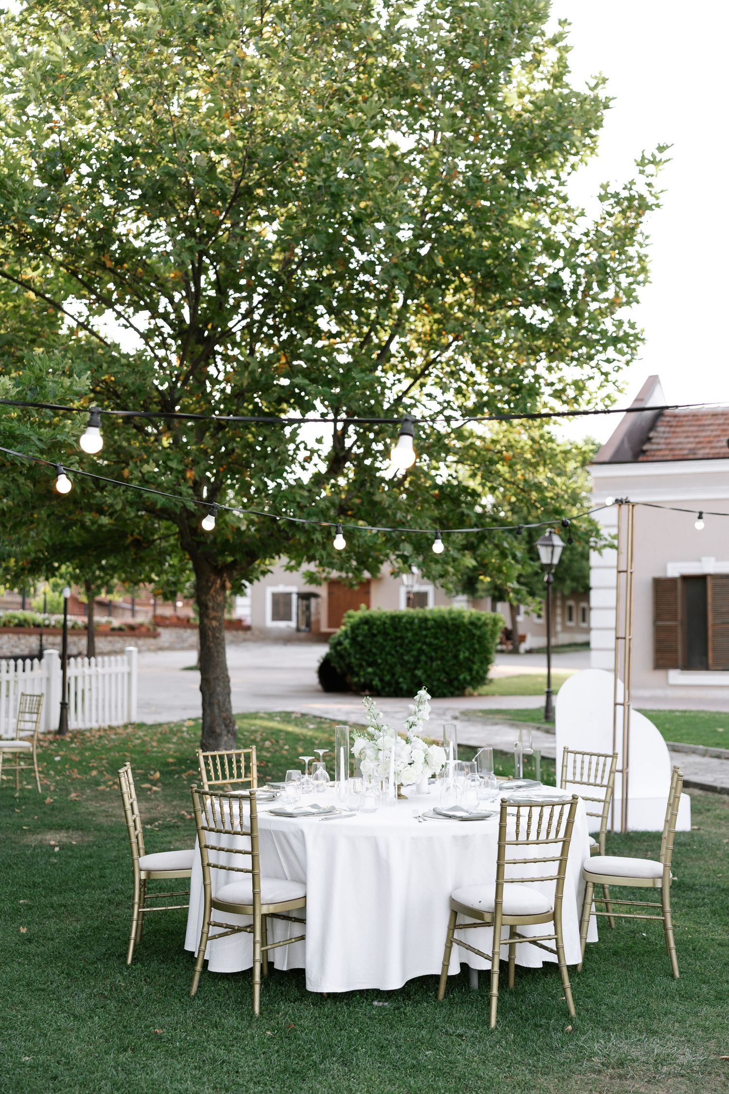 Elegant outdoor wedding reception table with white linens and gold chiavari chairs set beneath string lights and trees.