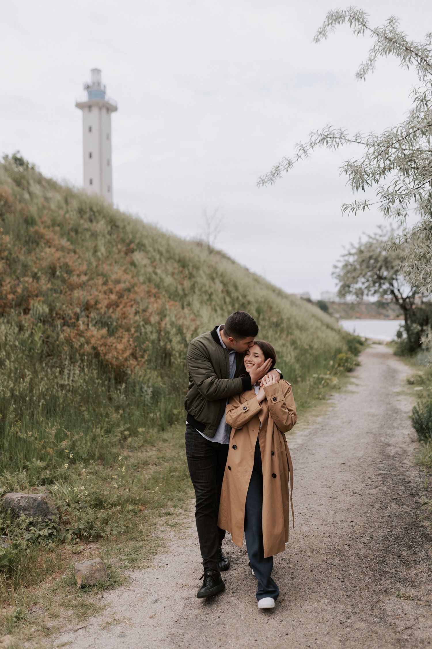 Couple sharing an intimate moment on a path with a lighthouse in the background.