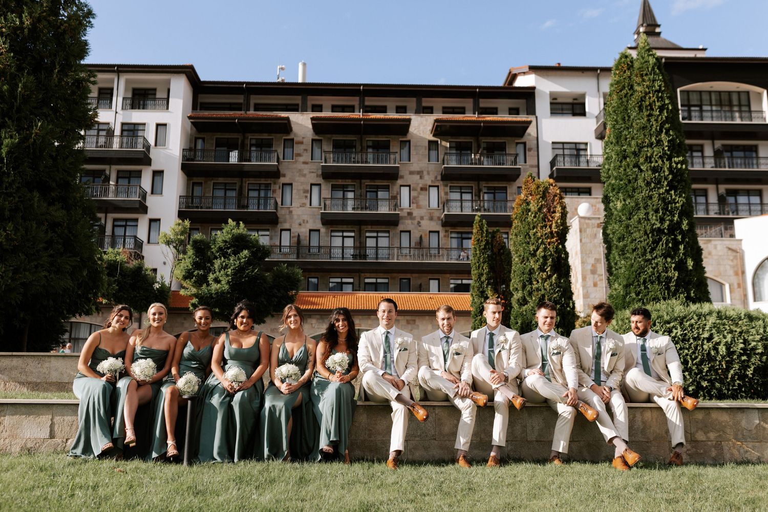 Wedding party in sage green and beige sits casually on stone steps at resort.