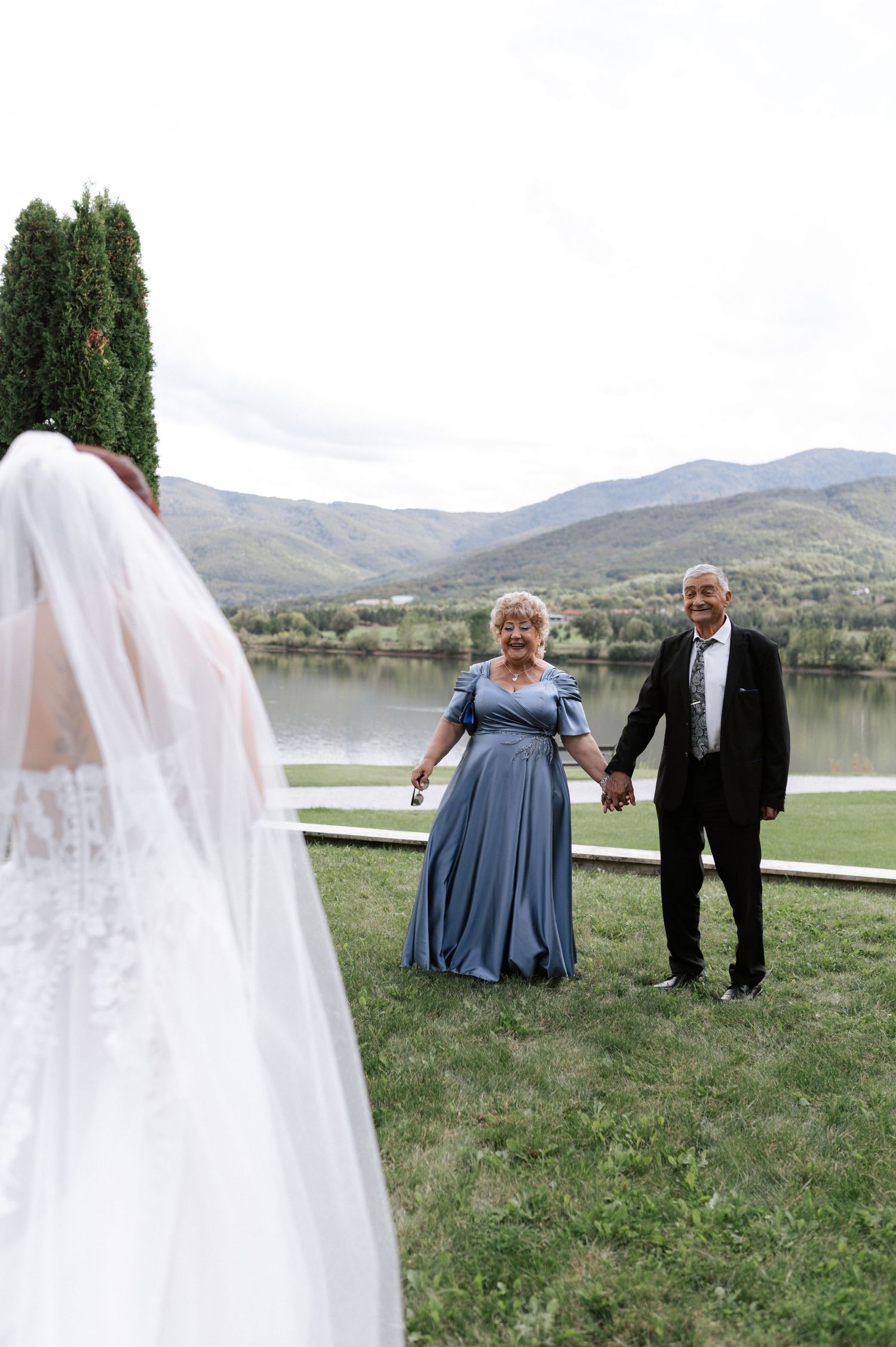 A wedding gathering by a serene lake with mountains in the background and a flowing white veil in the foreground.