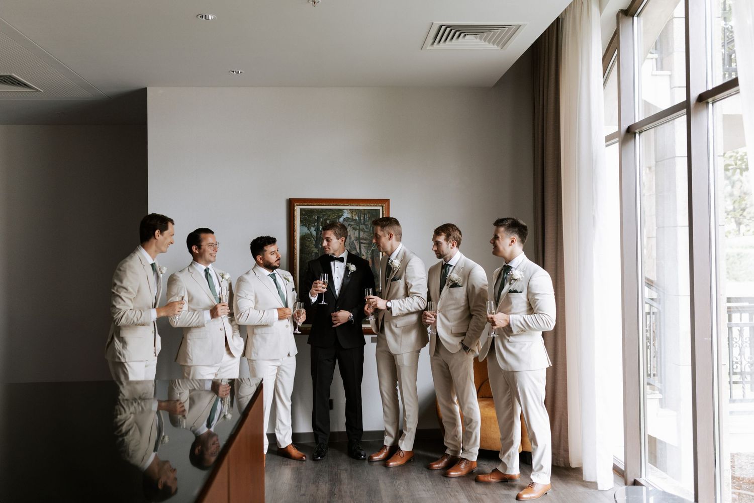 Groomsmen in light colored suits stand with groom in black tuxedo by large windows.