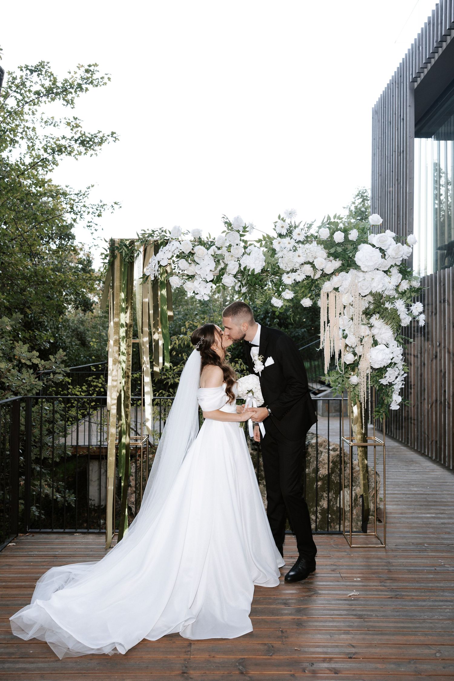 Intimate wedding moment captured under luxurious white floral arch installation.