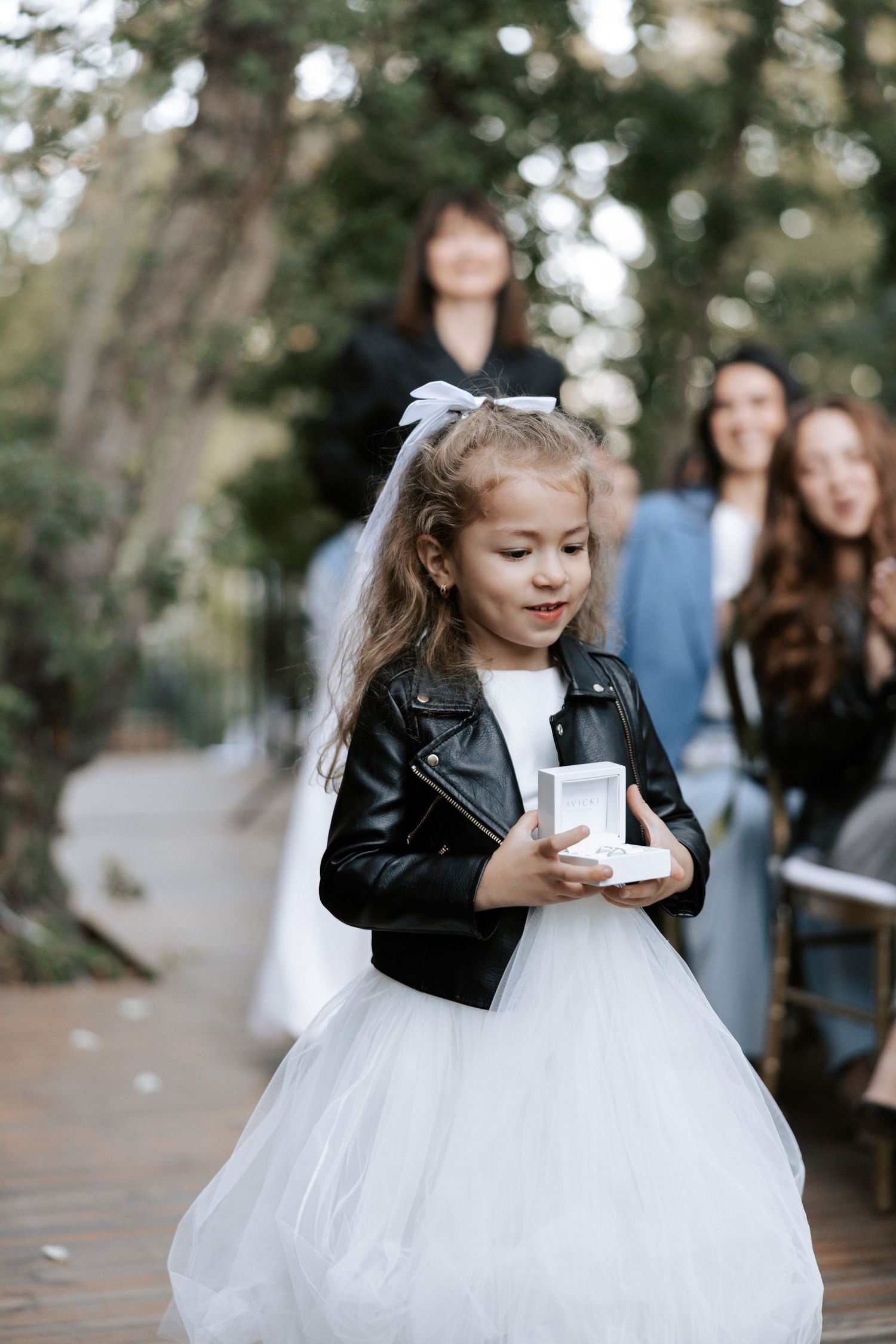 Young flower girl in white dress and leather jacket holds a small box at outdoor wedding.