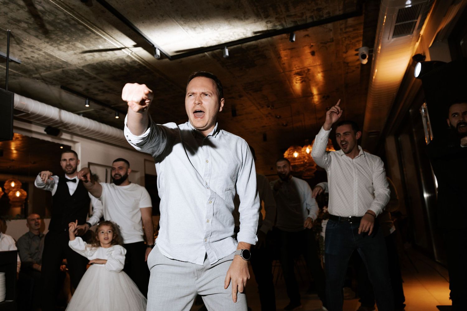 A person in a light colored suit jumps enthusiastically while dancing at an indoor event.