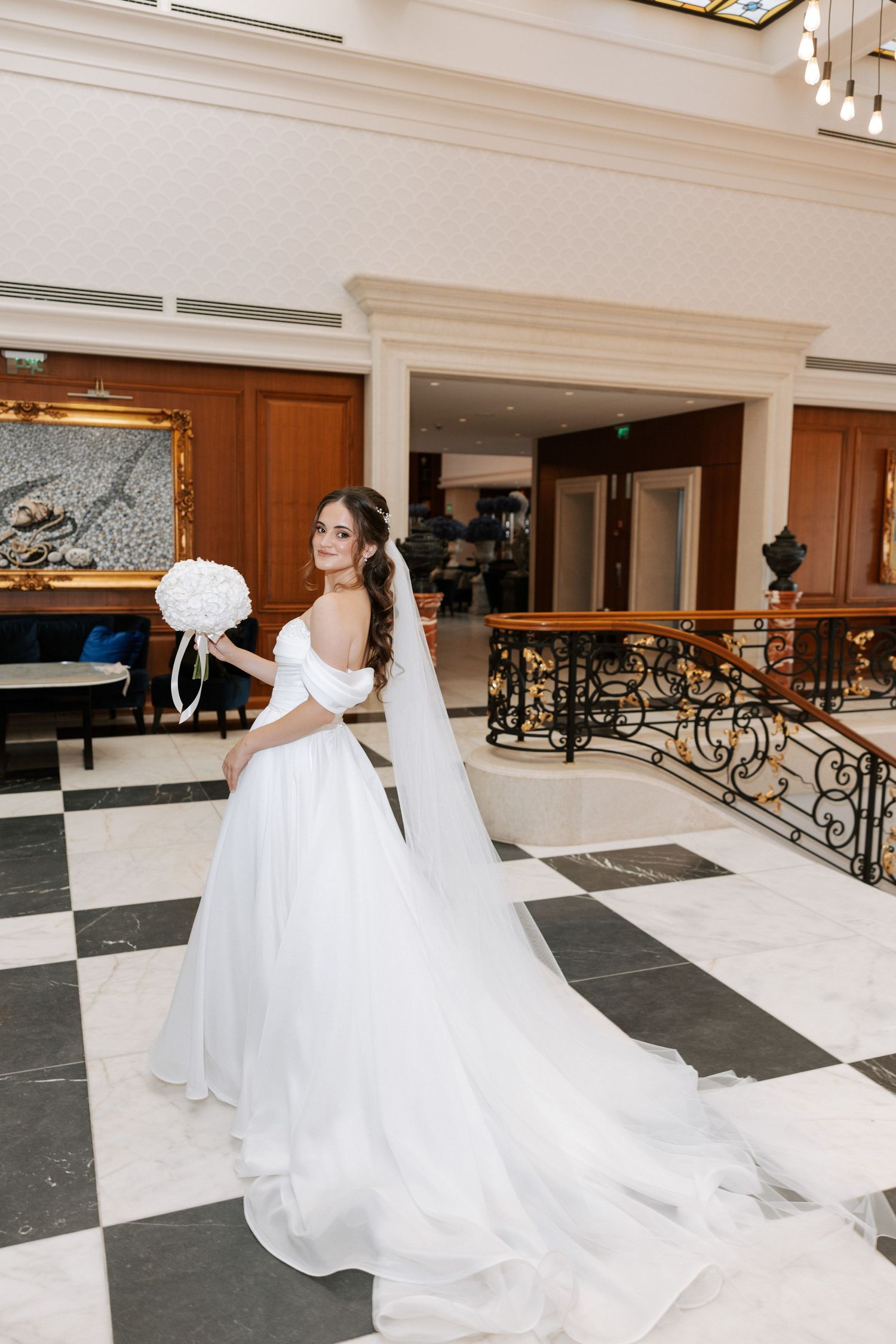 Bride in flowing white gown poses with bouquet on black and white checkered floor near ornate railing.
