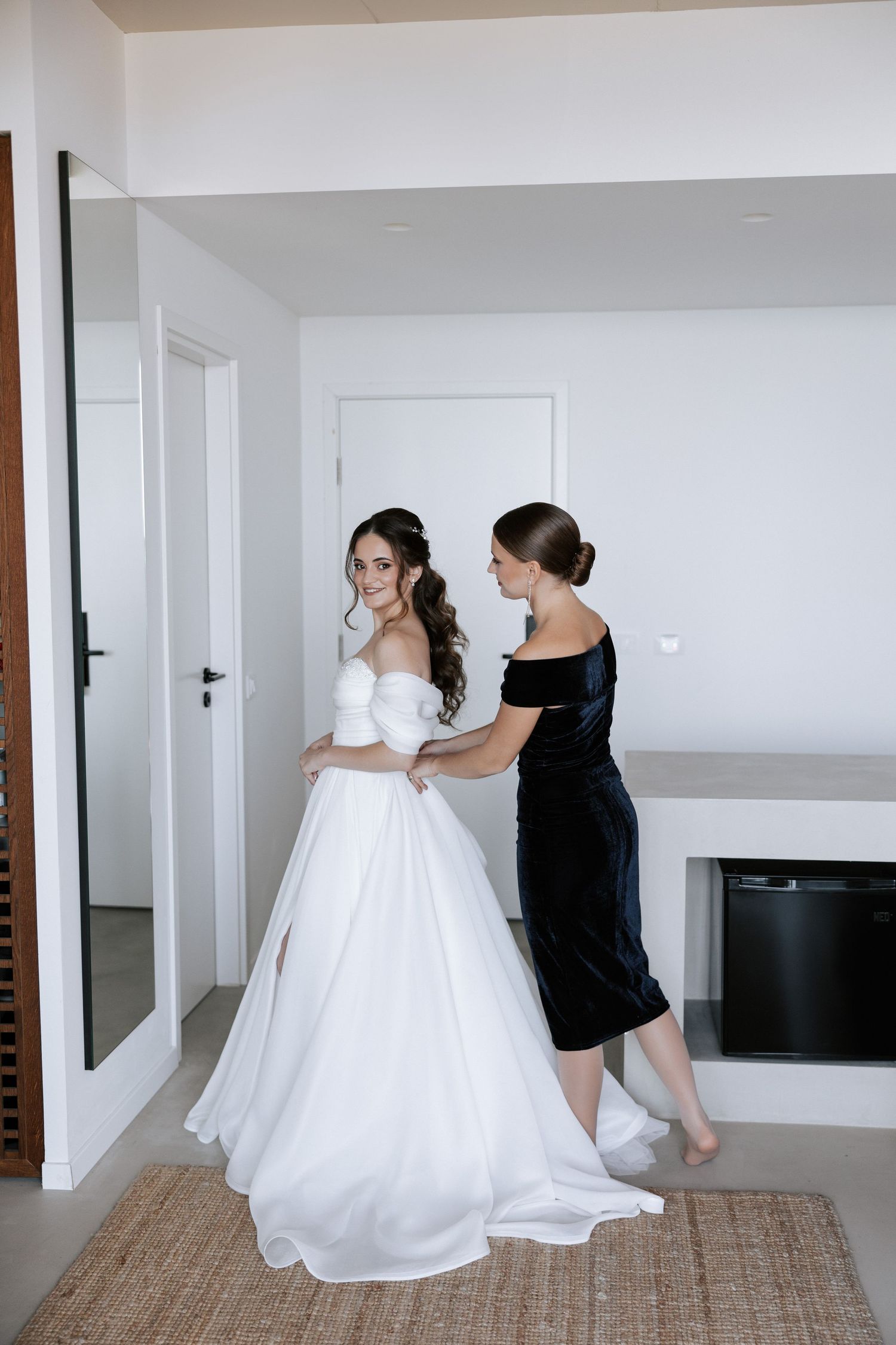 Woman in white wedding gown being helped with final dress adjustments by attendant in black dress.