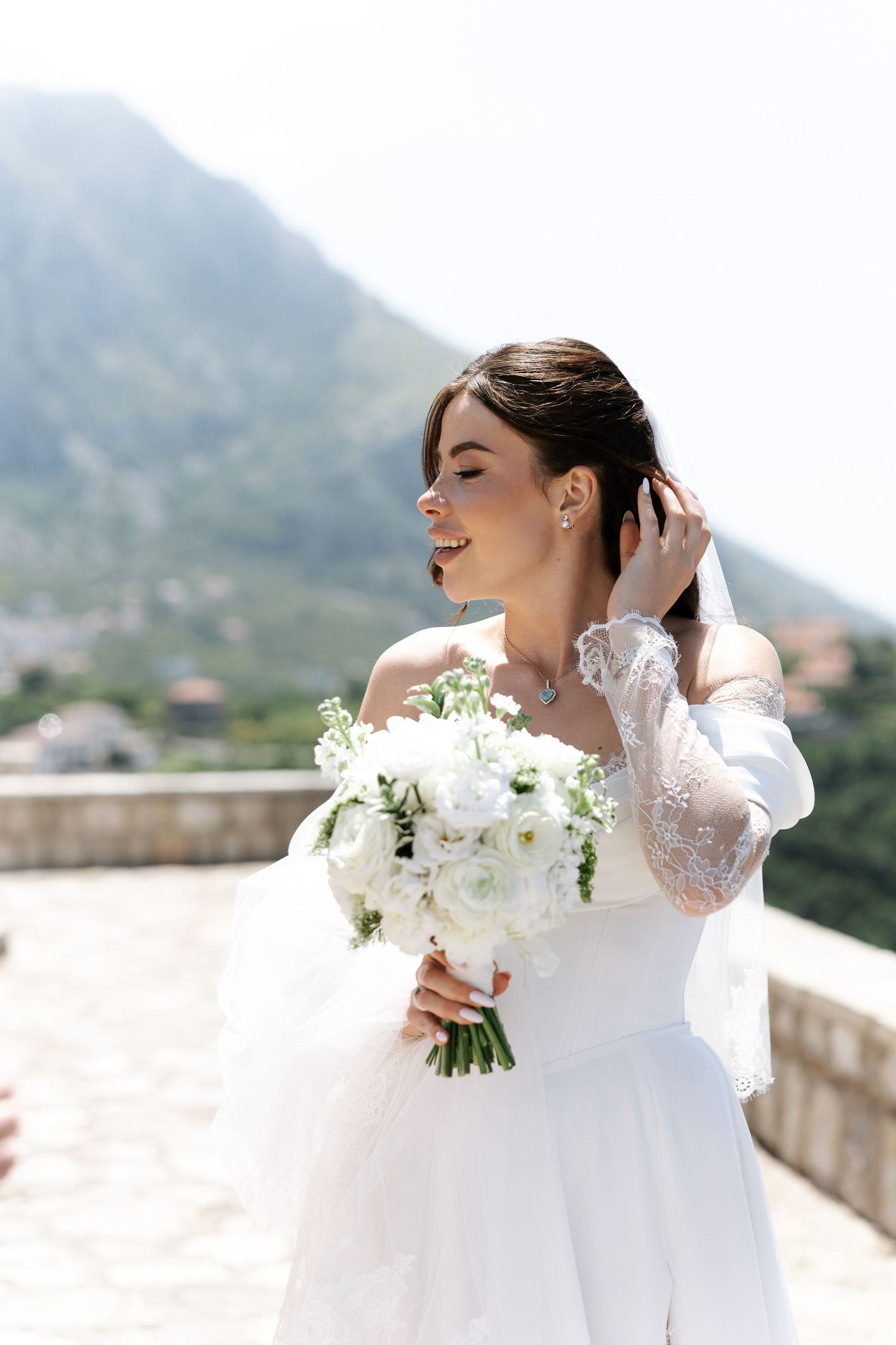 A bride in a white lace gown holds a bouquet of white flowers against a mountainous Mediterranean backdrop.
