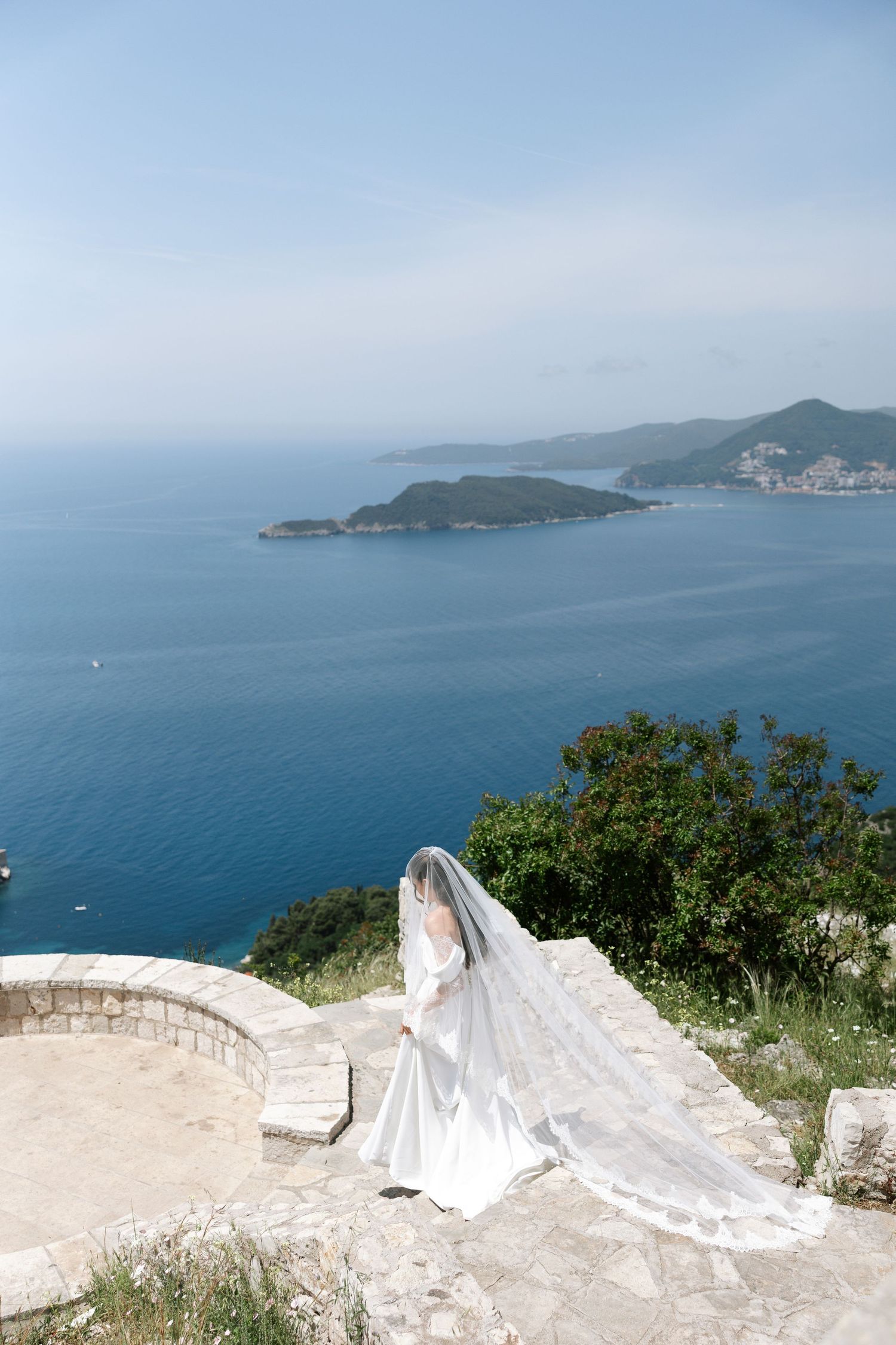 A flowing white wedding dress billows in the wind against a scenic coastal mountain backdrop overlooking blue ocean waters.