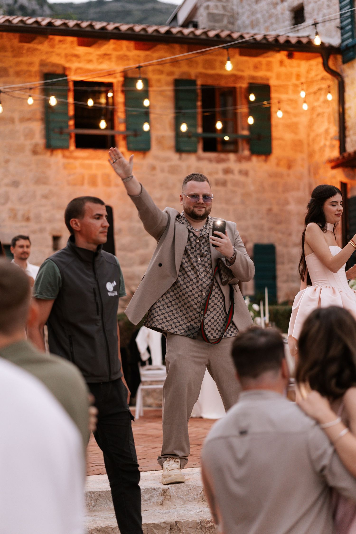 Wedding guests dance and celebrate at an outdoor reception with string lights and stone walls in the background.
