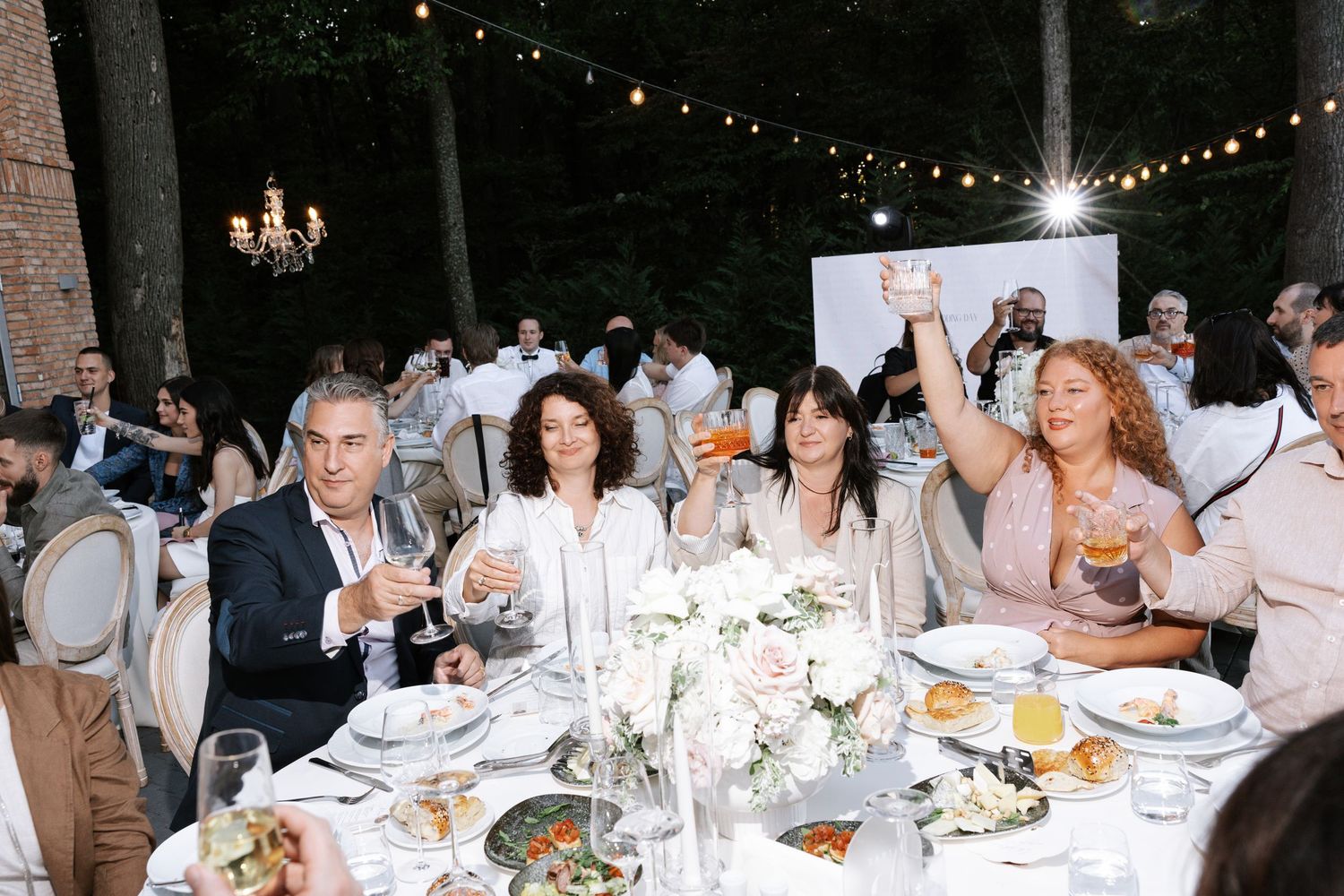Evening wedding reception dinner under string lights with guests at long tables.