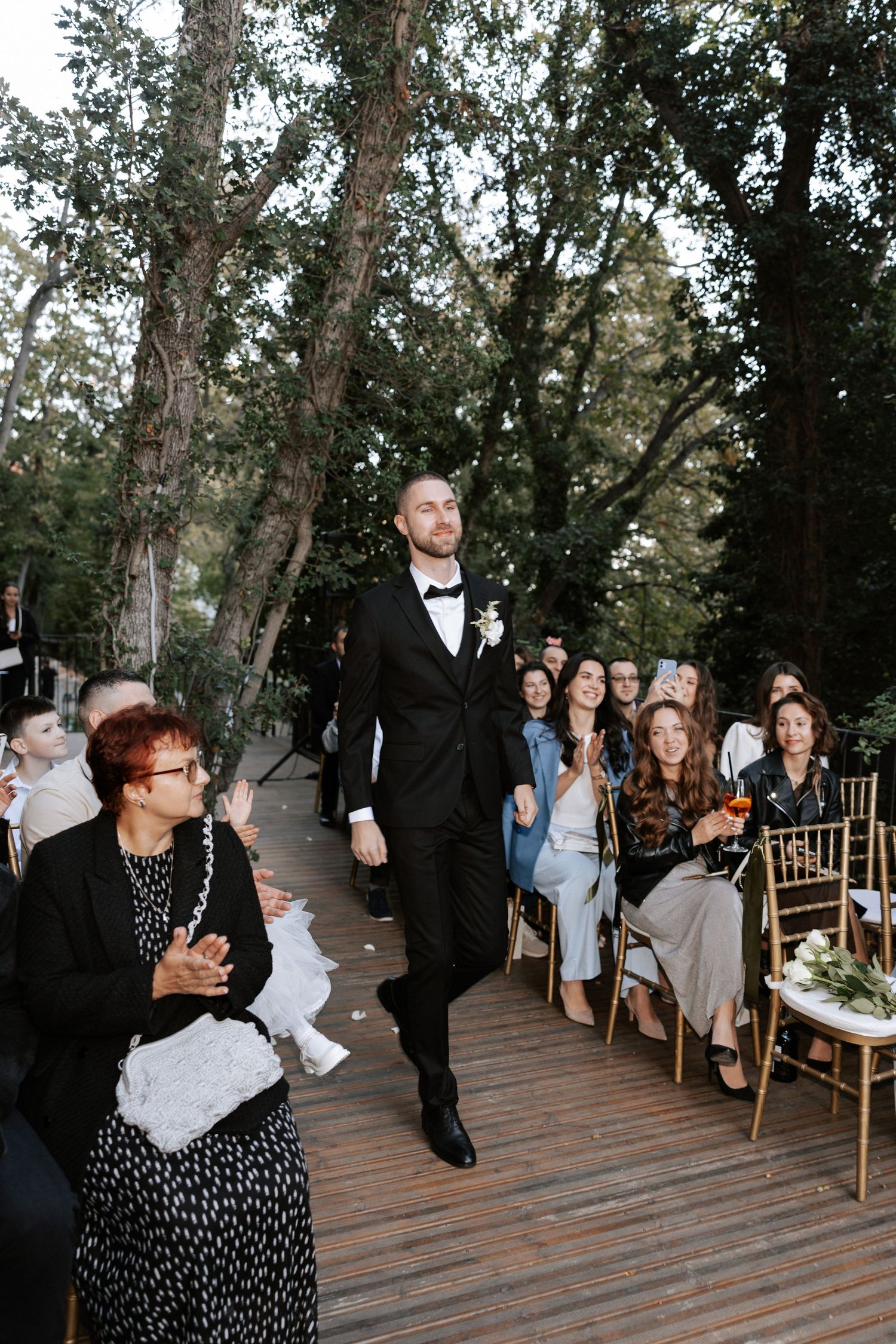 A formal ceremony processional takes place on a wooden deck with trees in the background.