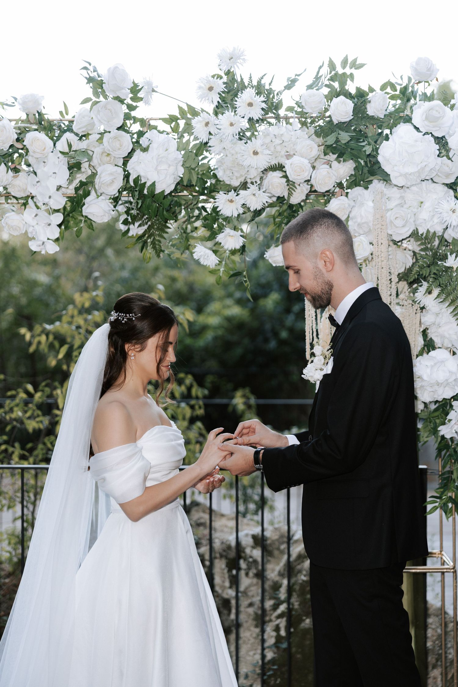 A beautiful wedding ceremony beneath a floral arch of white flowers and greenery on a wooden deck.