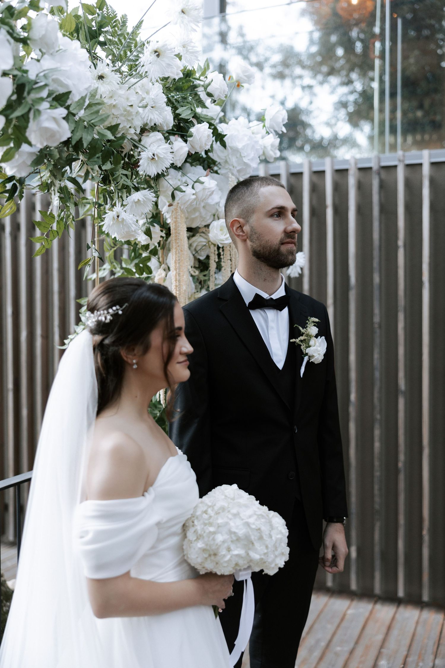 An elegant wedding couple stands under a white floral arch during their outdoor ceremony.