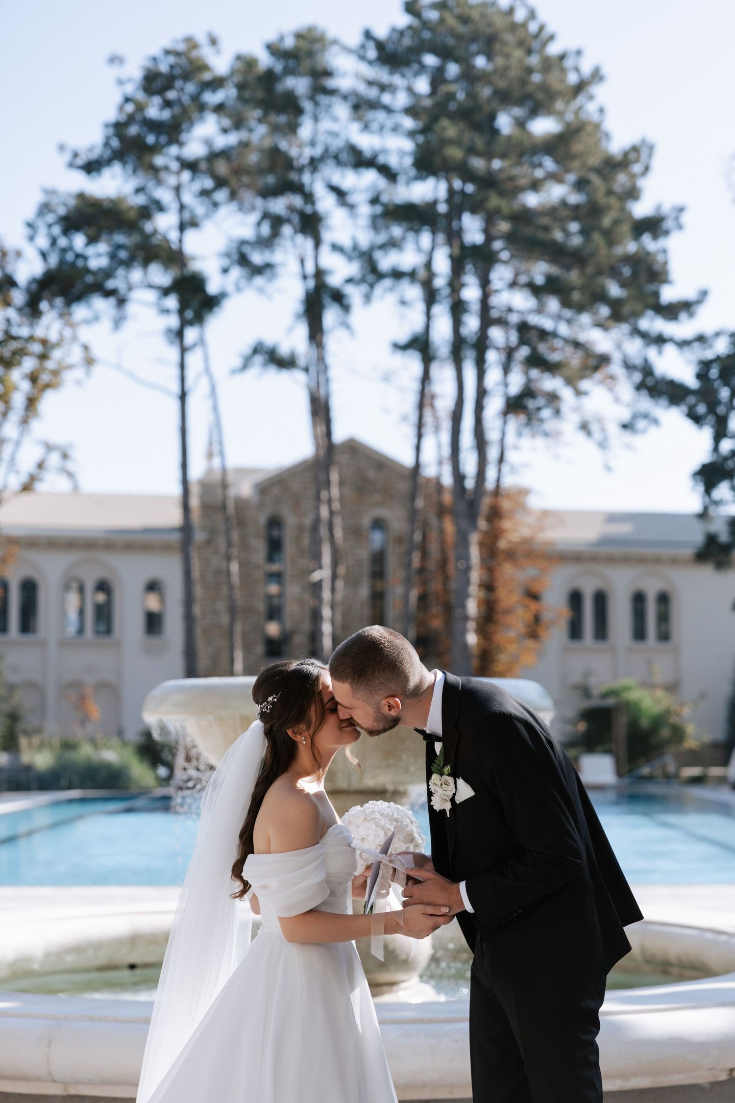 Romantic moment between newlyweds by an elegant fountain with tall pine trees.