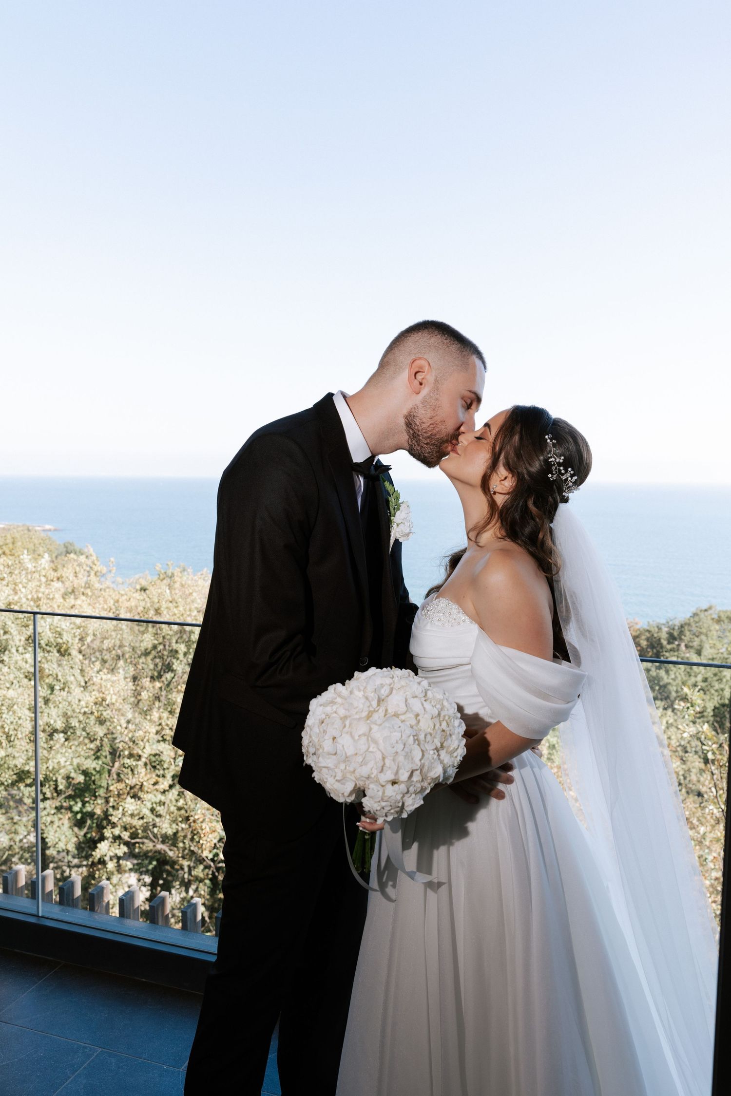 A romantic wedding couple shares a kiss on an oceanview balcony with a stunning white floral bouquet.