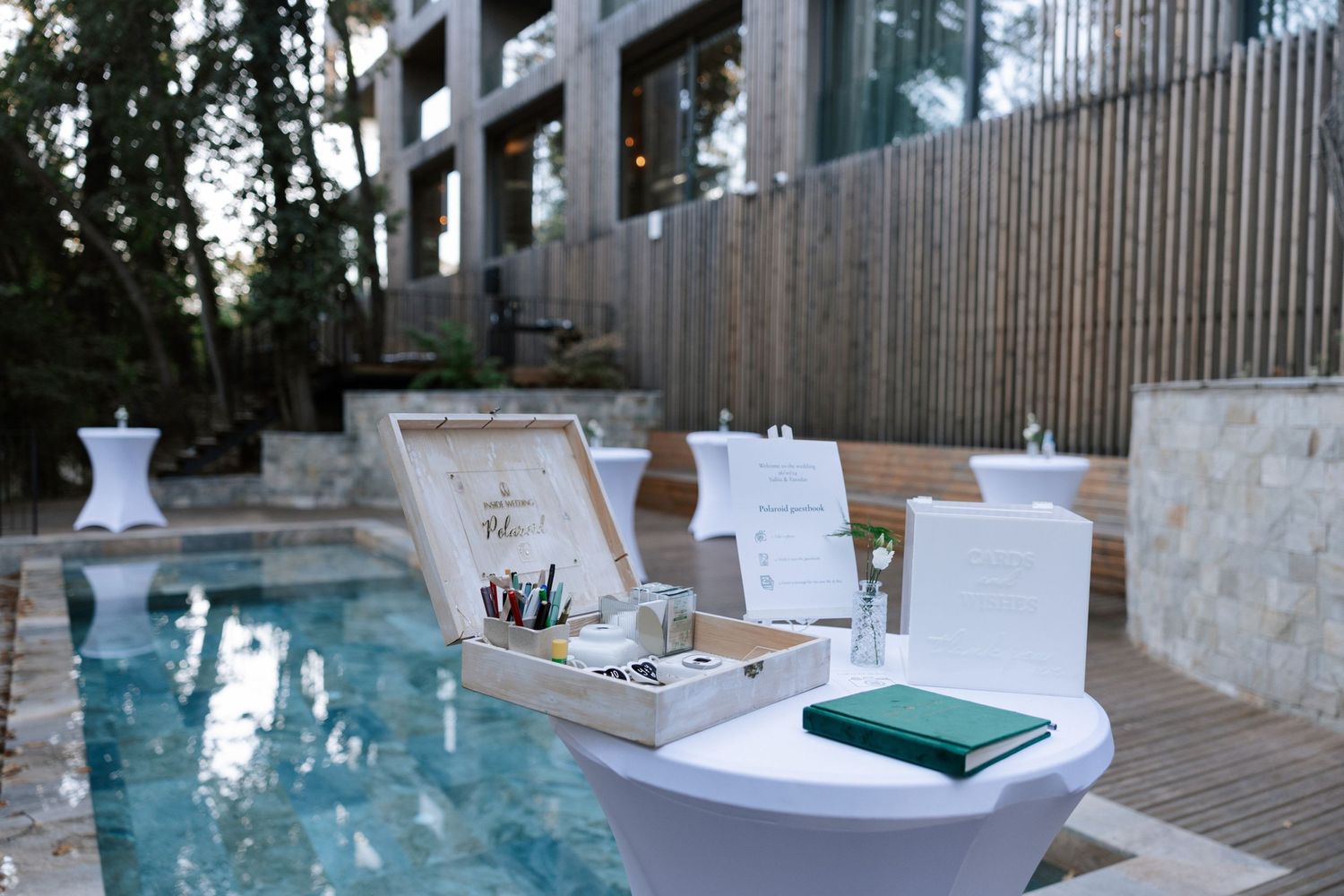 Modern poolside wedding setup with white decor and signage near contemporary building.