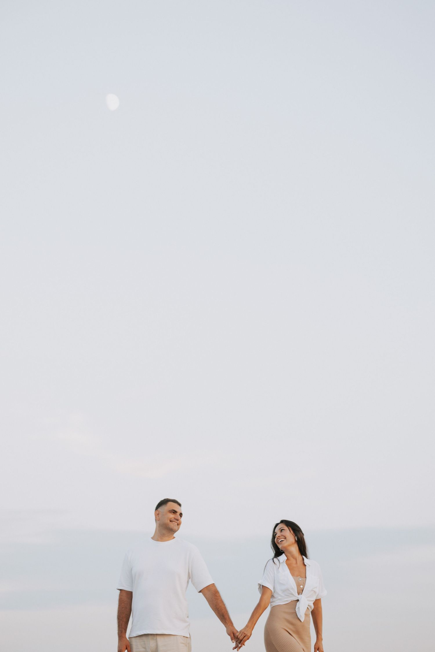 A couple holds hands against a misty sky with a faint moon visible above.