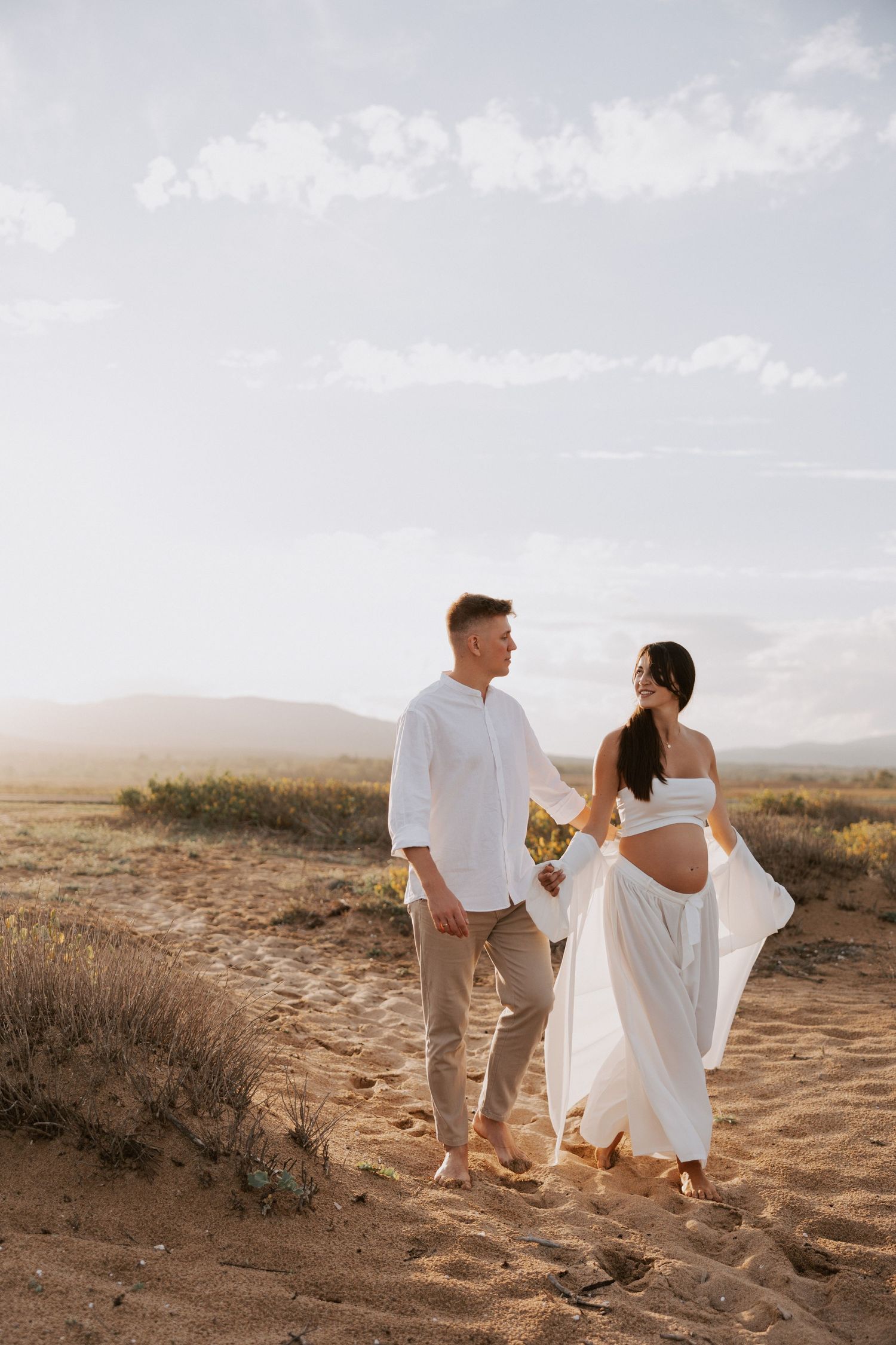 Couple in white attire holds hands while walking on desert path with mountains in background.