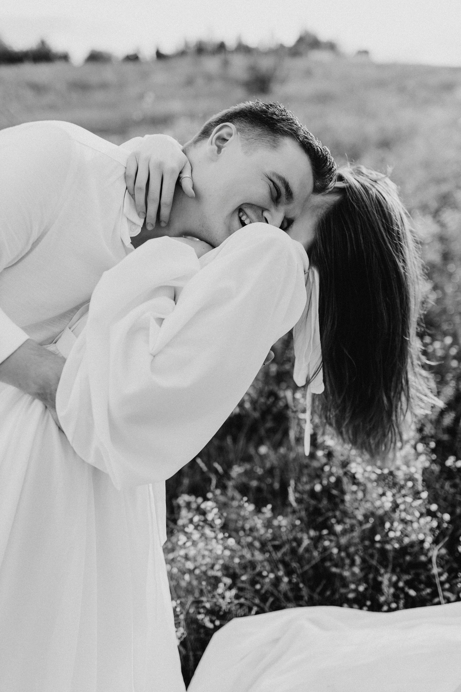 Romantic black and white portrait of couple embracing in a meadow wearing white flowing outfits.