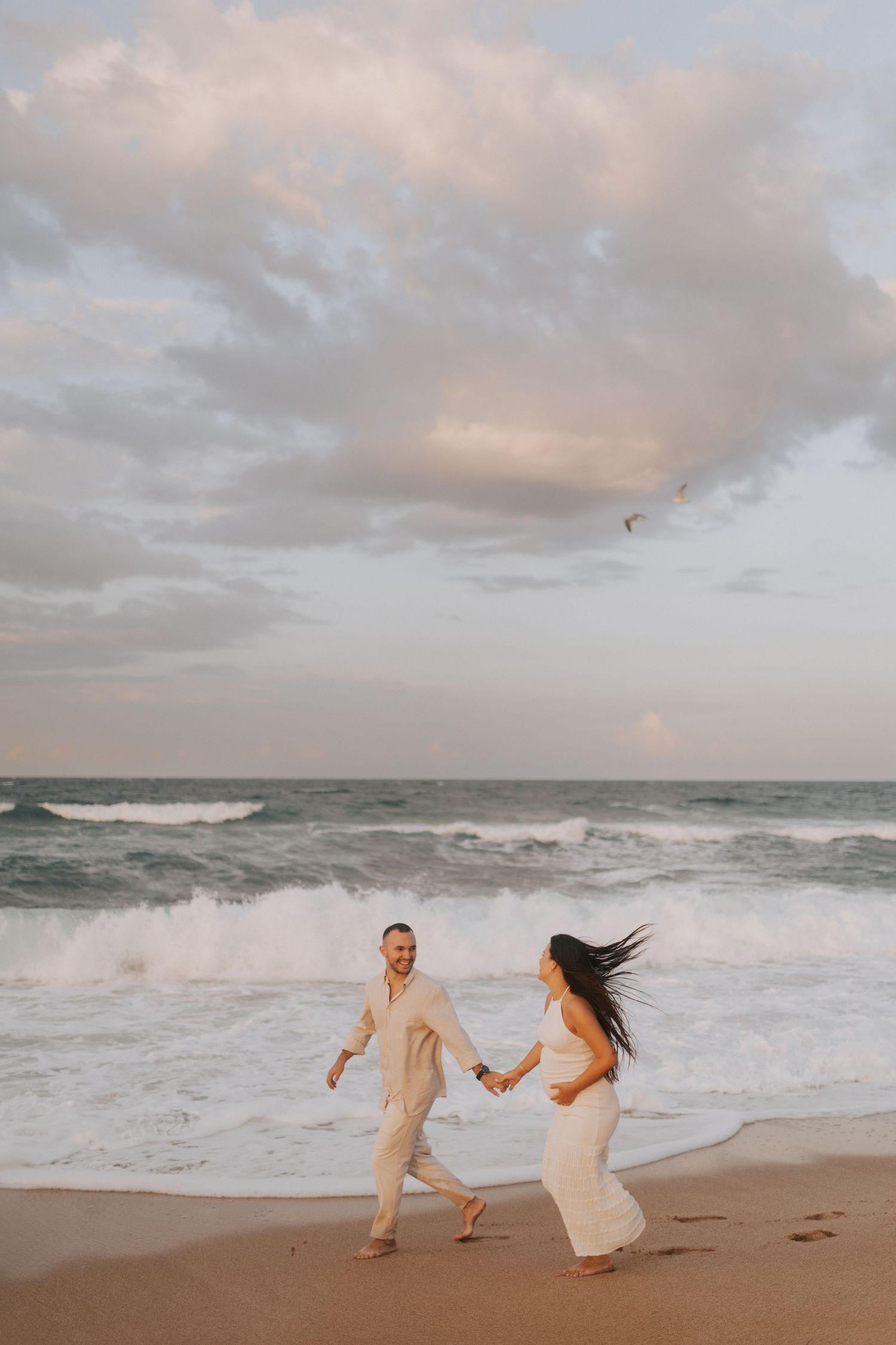 A couple runs playfully along the beach with crashing waves and cloudy skies in the background.