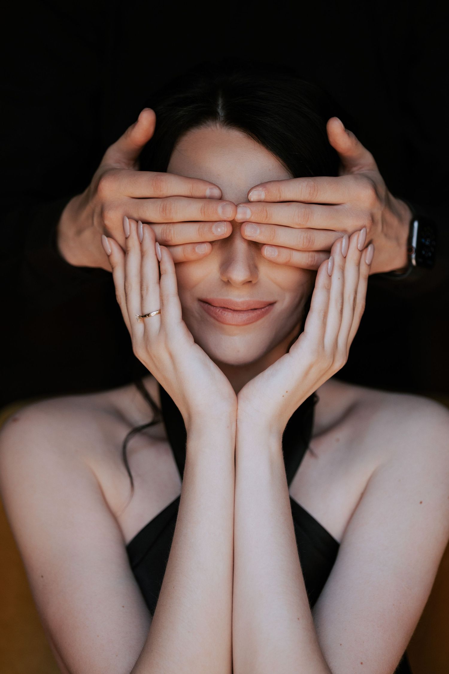 Hands cover eyes in artistic portrait showing gentle smile and bare shoulders.