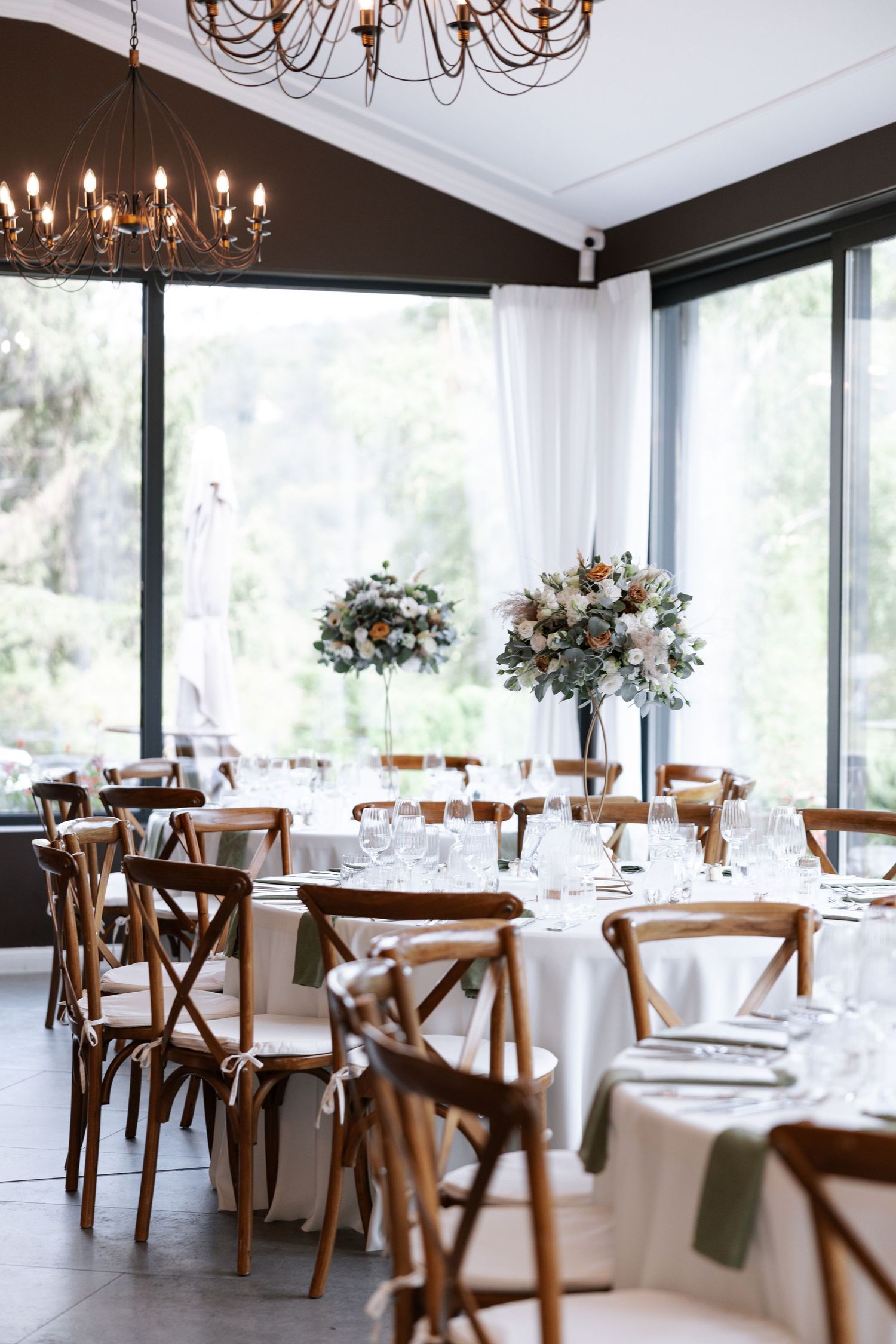 Elegant wedding reception venue with white tablecloths, wooden cross-back chairs, and crystal chandeliers in natural light.